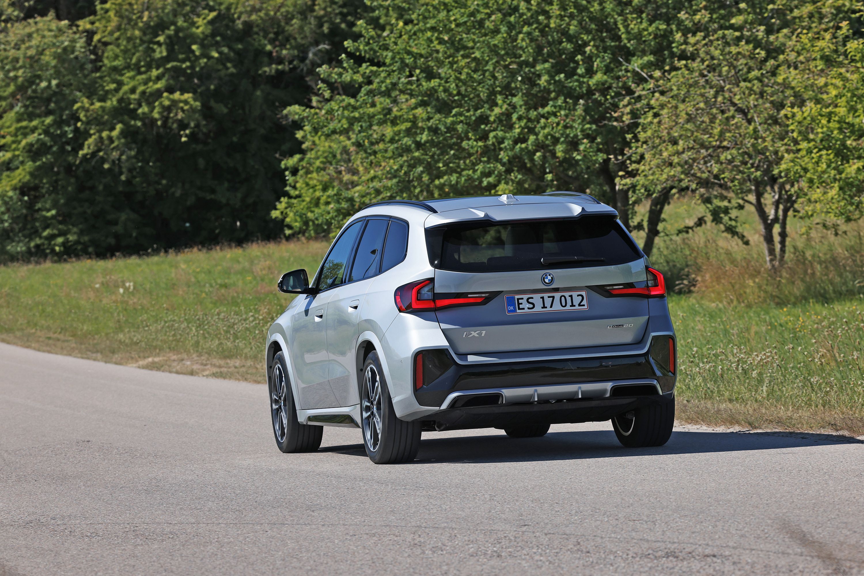 Den faceliftede udgave af BMW iX1 eDrive Supercharged fra 2026 set bagfra på en landevej med grønne træer i baggrunden. 