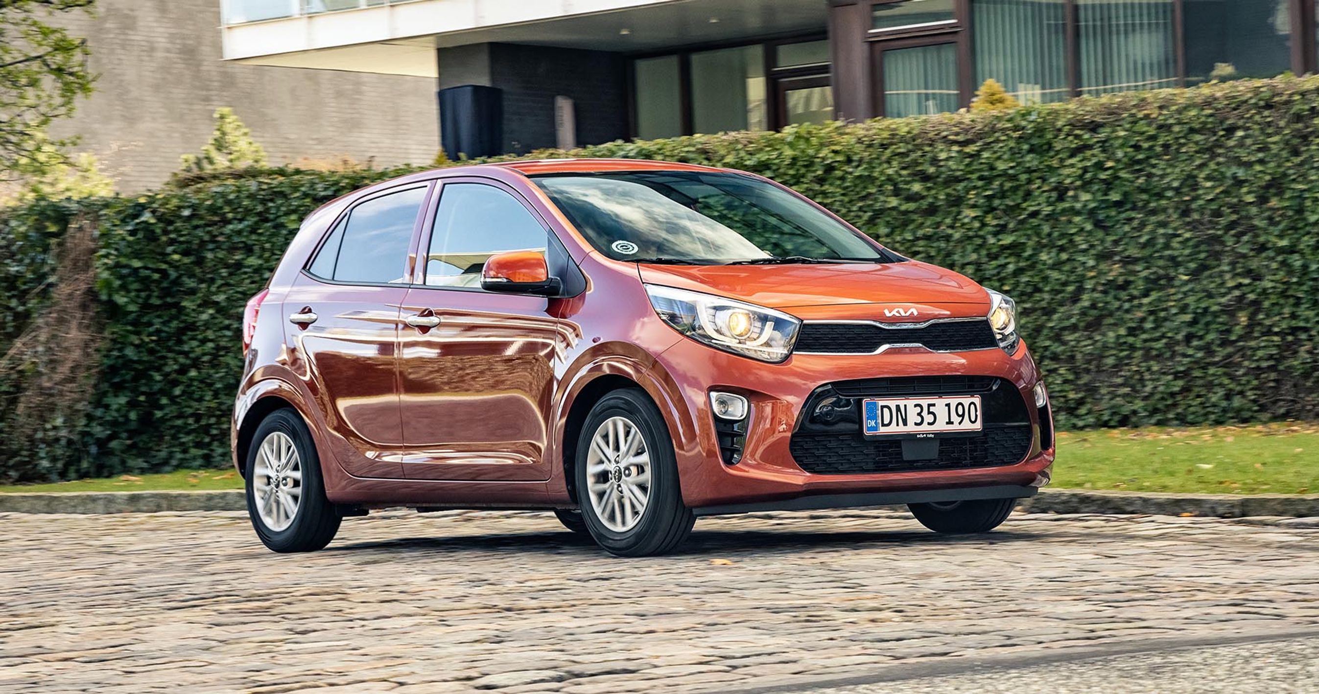 Orange Kia Picanto kører på brosten. I baggrunden en grøn hæk.