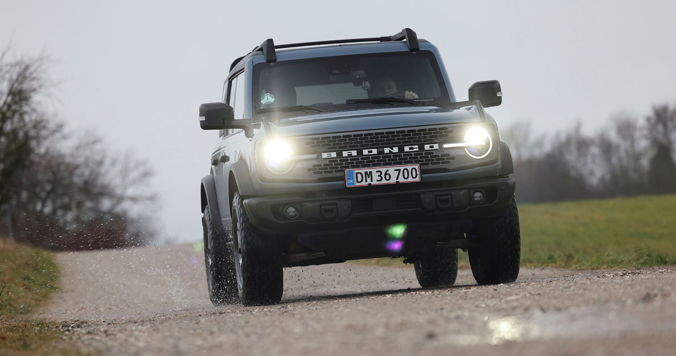 En Ford Bronco kører på en grusvej i tåge med forlygterne tændt