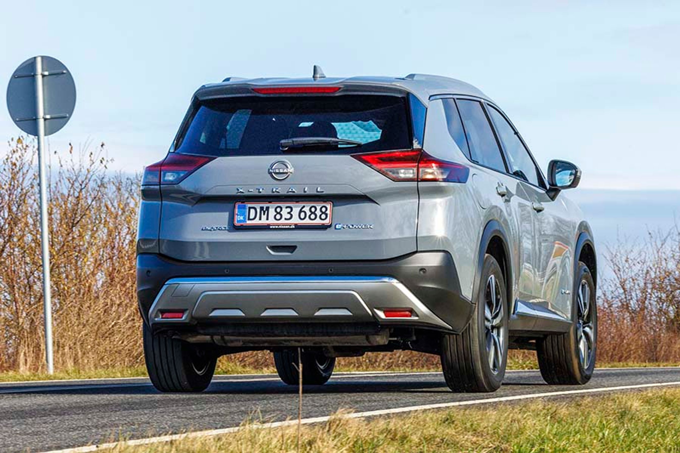 Nissan X-Trail set bagfra, mens den kører på en landevej med buske og et skilt i baggrunden
