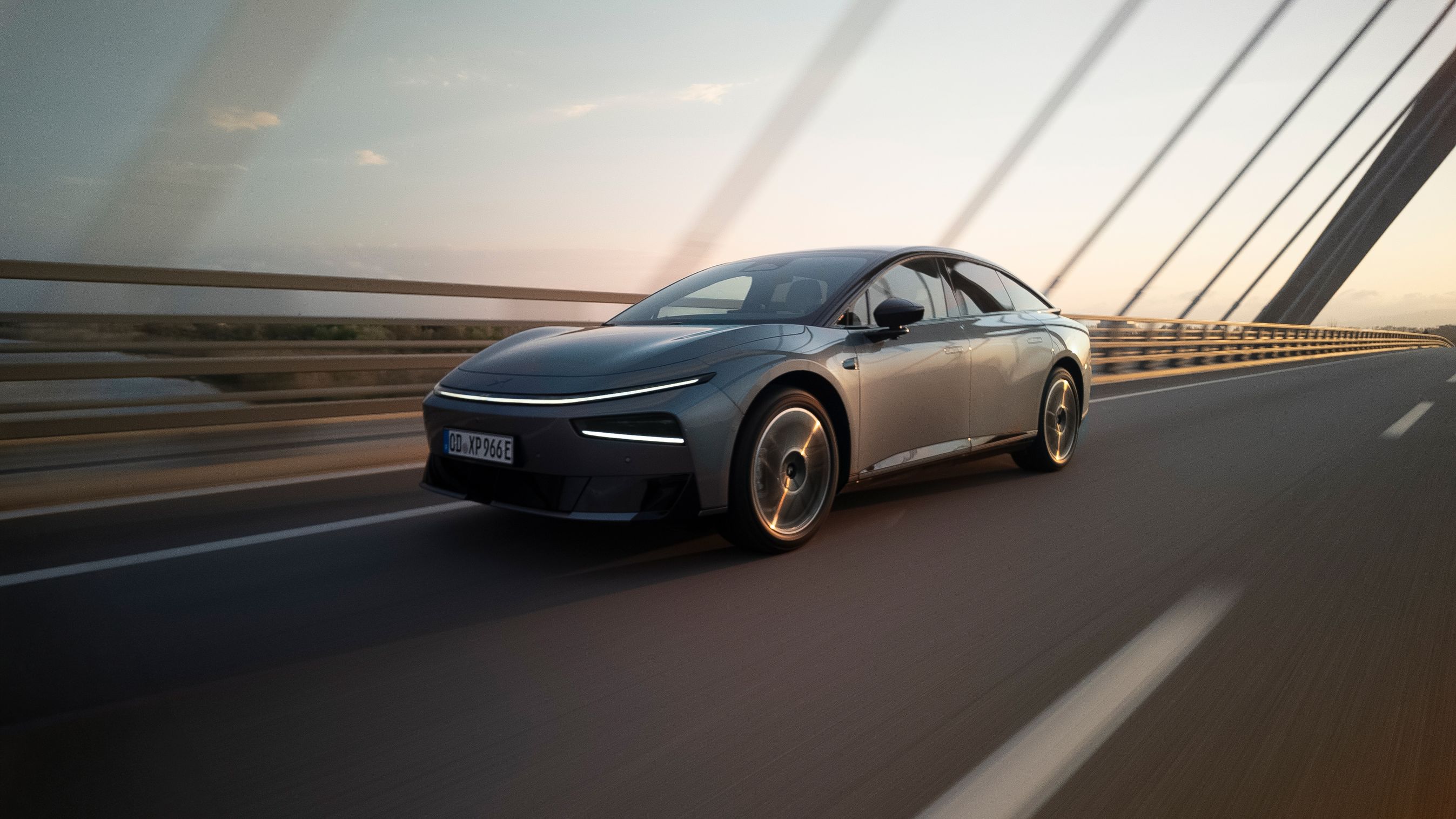 Xpeng P7+ set skråt forfra, mens den kører på bro i solnedgang