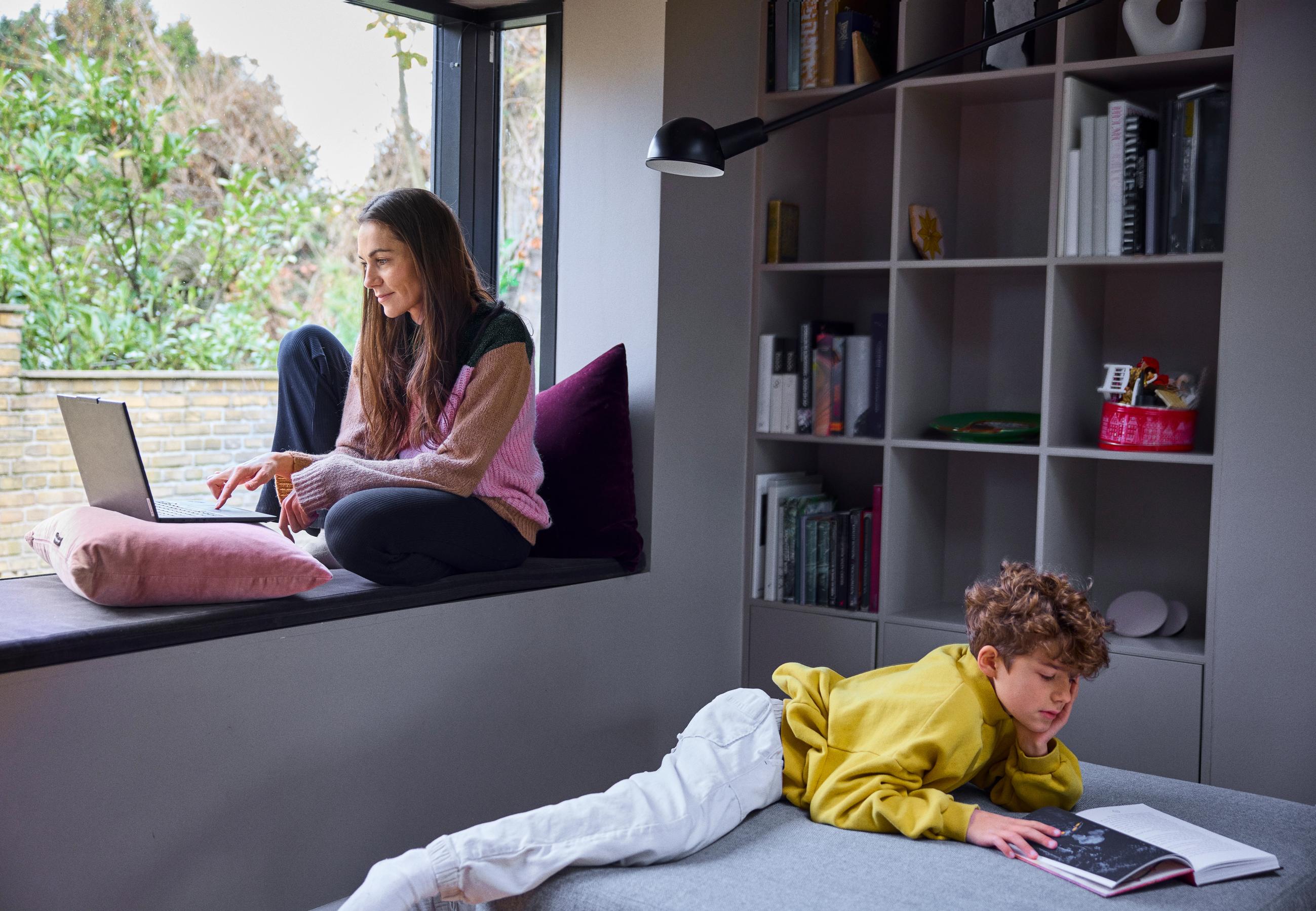 mor sidder i vinduskarm med hendes computer og drengen læser bog i deres stue