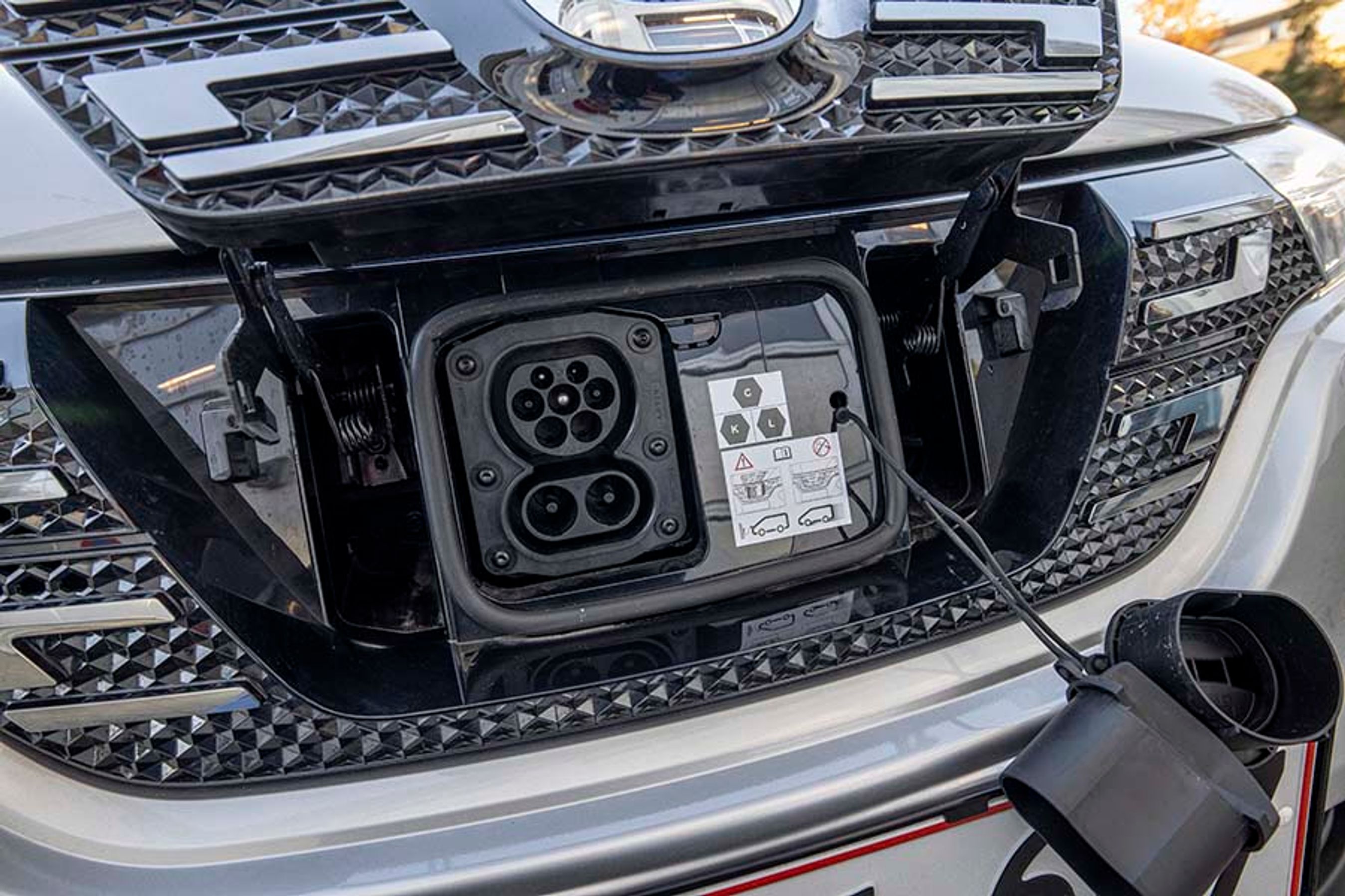 Ladestikket er placeret i fronten af bilen, og man skal selv fjerne gummi-propperne, når stikket skal bruges. Bilen har som standard en et-faset AC-opladning med maksimalt 3,7 kW, men man kan for 5.000 kr. tilkøbe et DC-ladestik (nederst), og så kan bilen oplades med op til 30 kW.