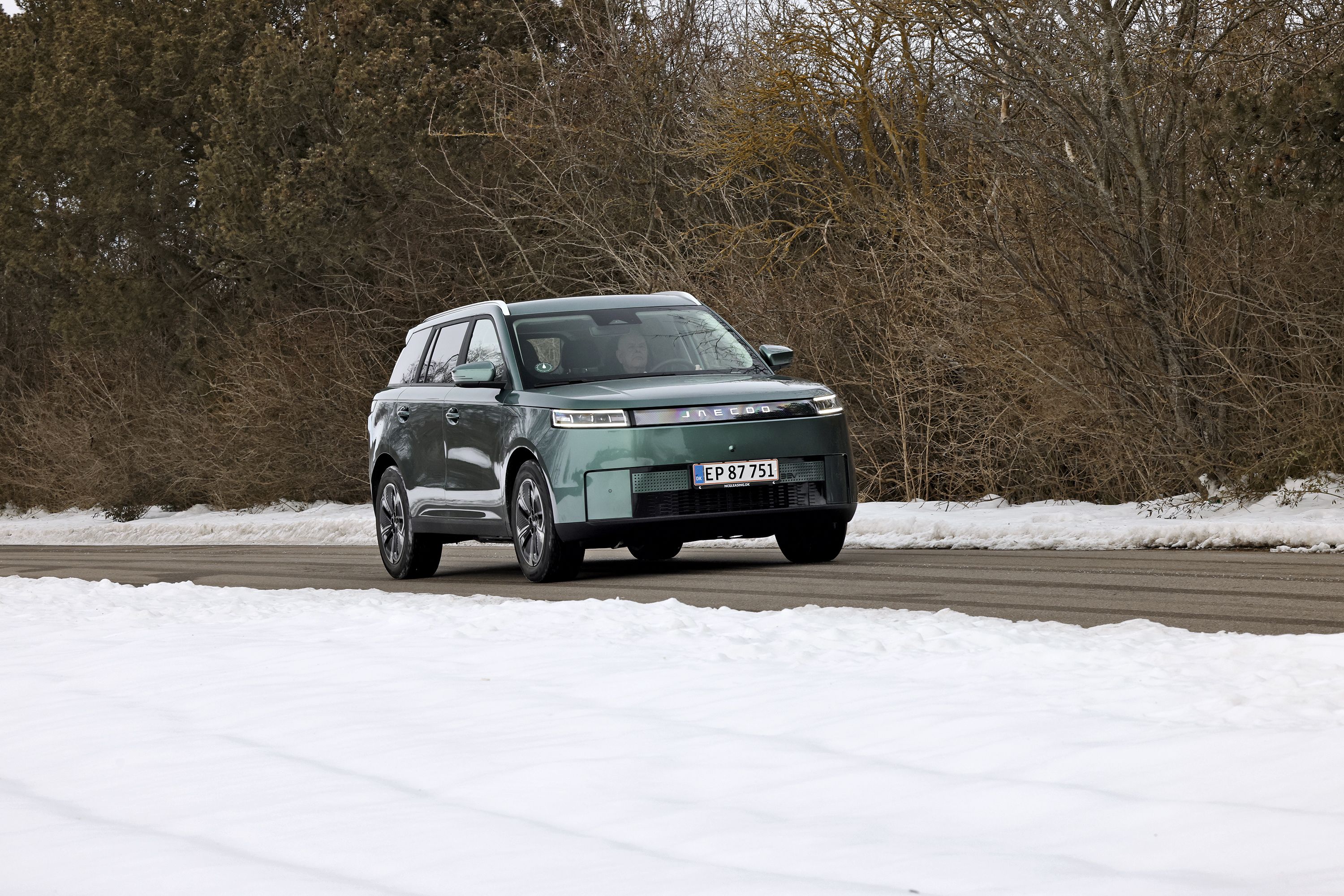 Grøn Jaecoo J5 kører på landevej. Der er sne i forgrunden og bare grene i baggrunden. 