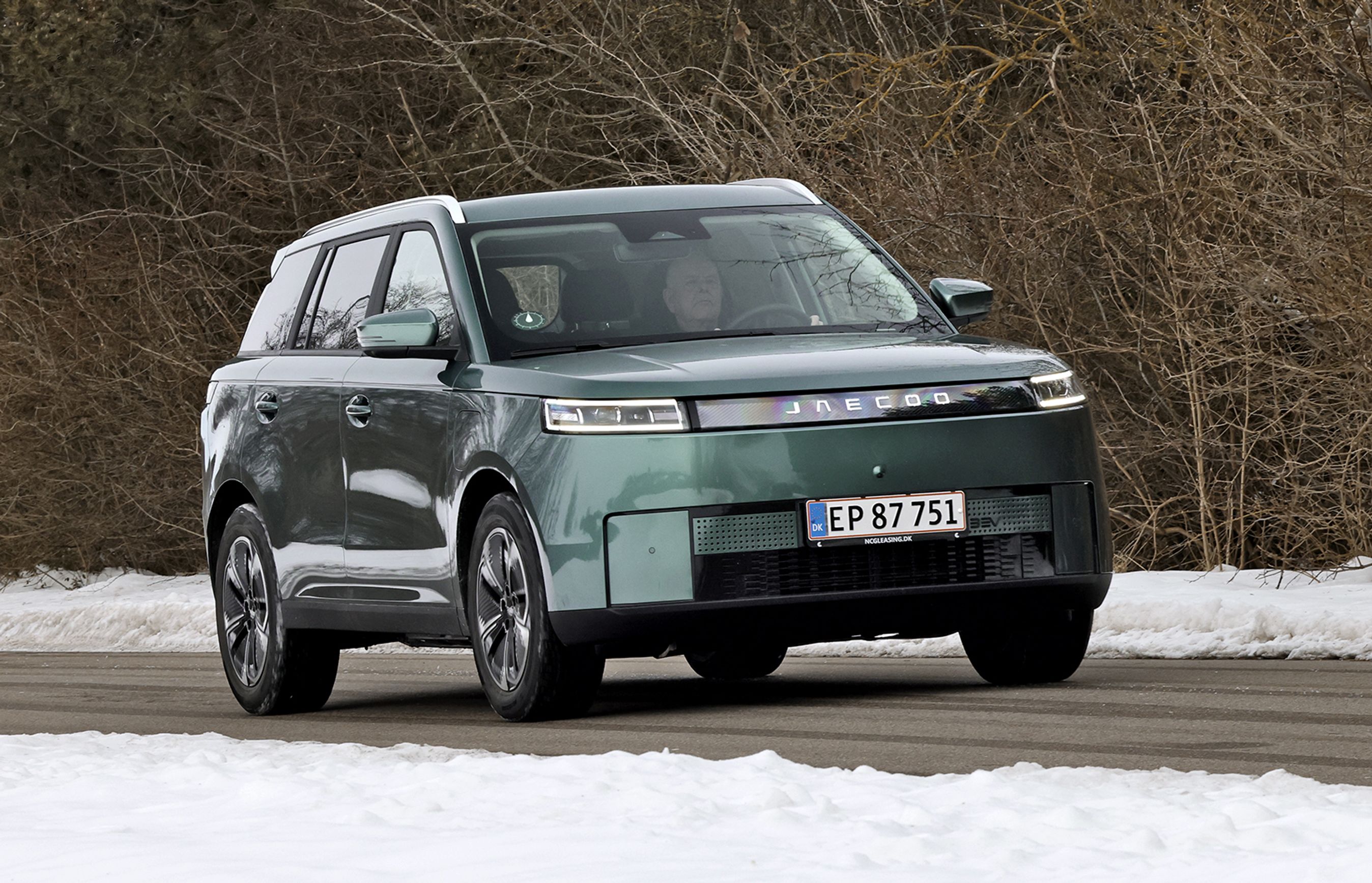 Grøn Jaecoo J5 kører på landevej. Der er sne i forgrunden og bare grene i baggrunden.