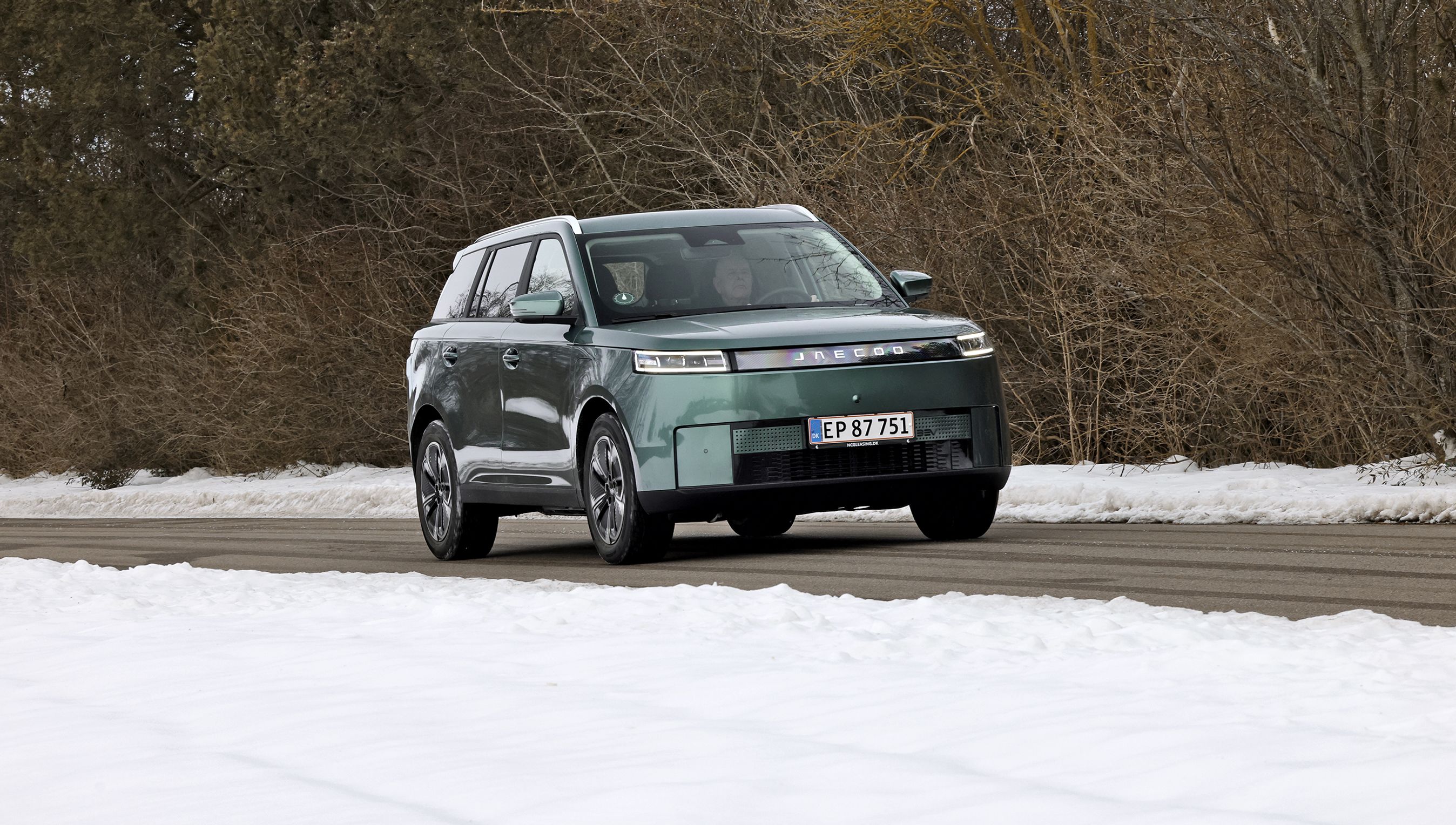 Grøn Jaecoo J5 kører på landevej. Der er sne i forgrunden og bare grene i baggrunden.