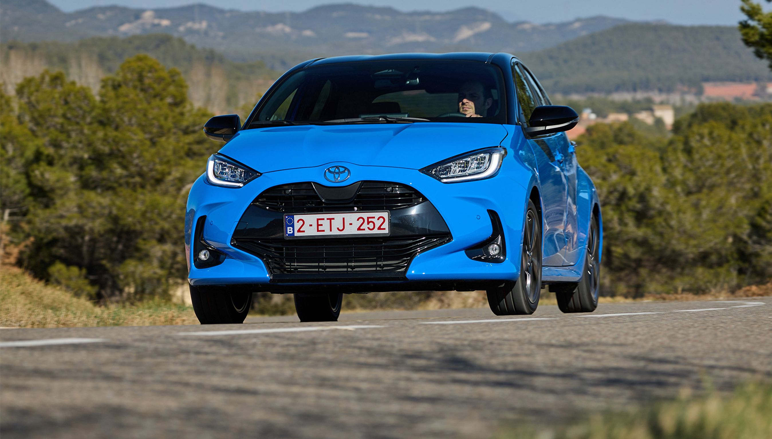 En blå Toyota Yaris kører på en vej med træer og bjerge i baggrunden