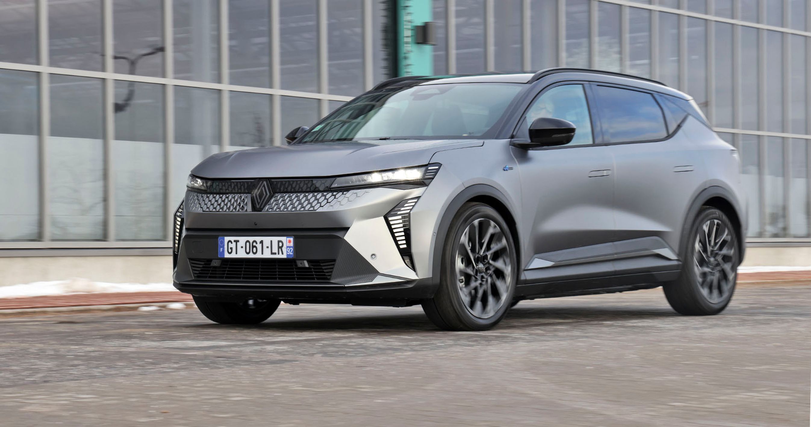 En mørkegrå Renault Scenic holder foran en bygning med mange vinduer