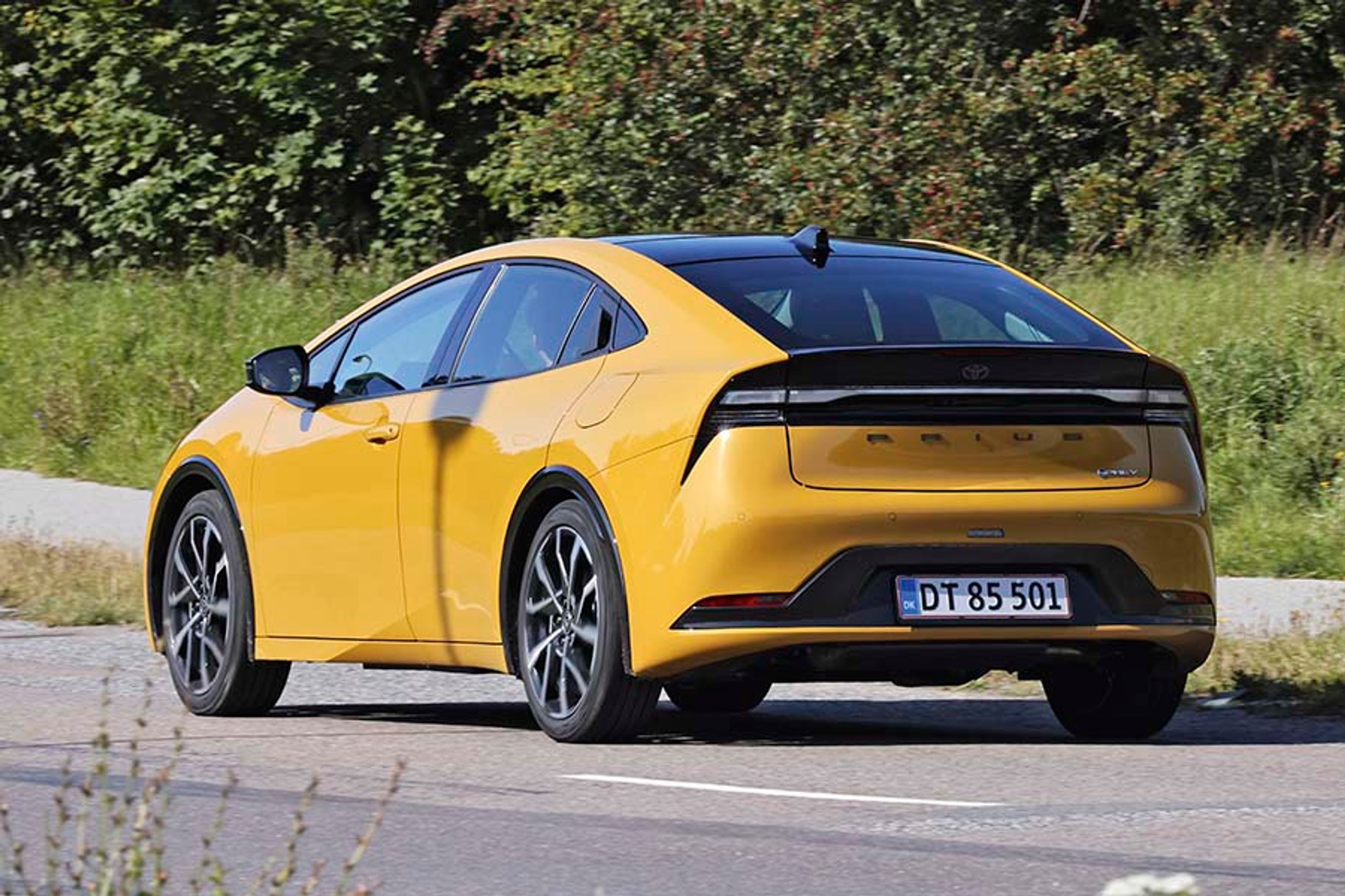 En gul Toyota Prius set bagfra kører på en vej med buske i baggrunden.