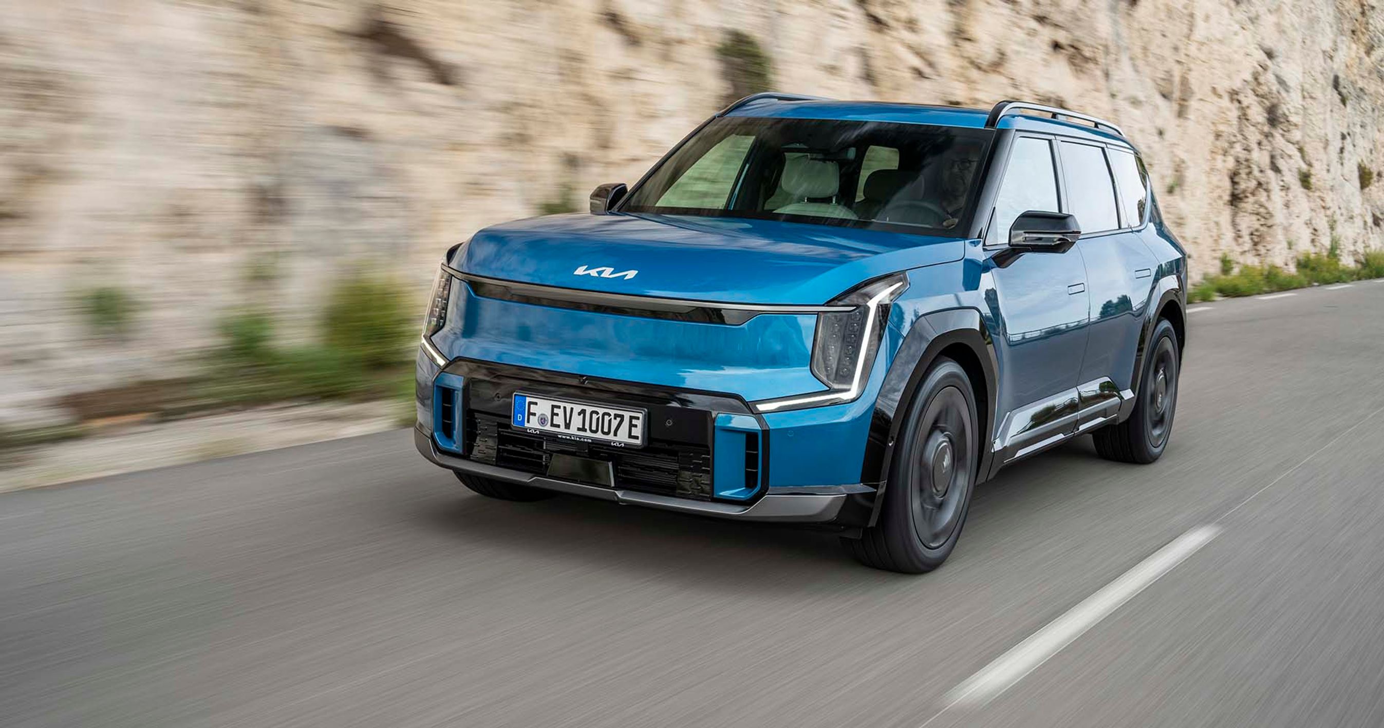 En blå Kia EV6 elbil kører på en bjergvej med klipper i baggrunden