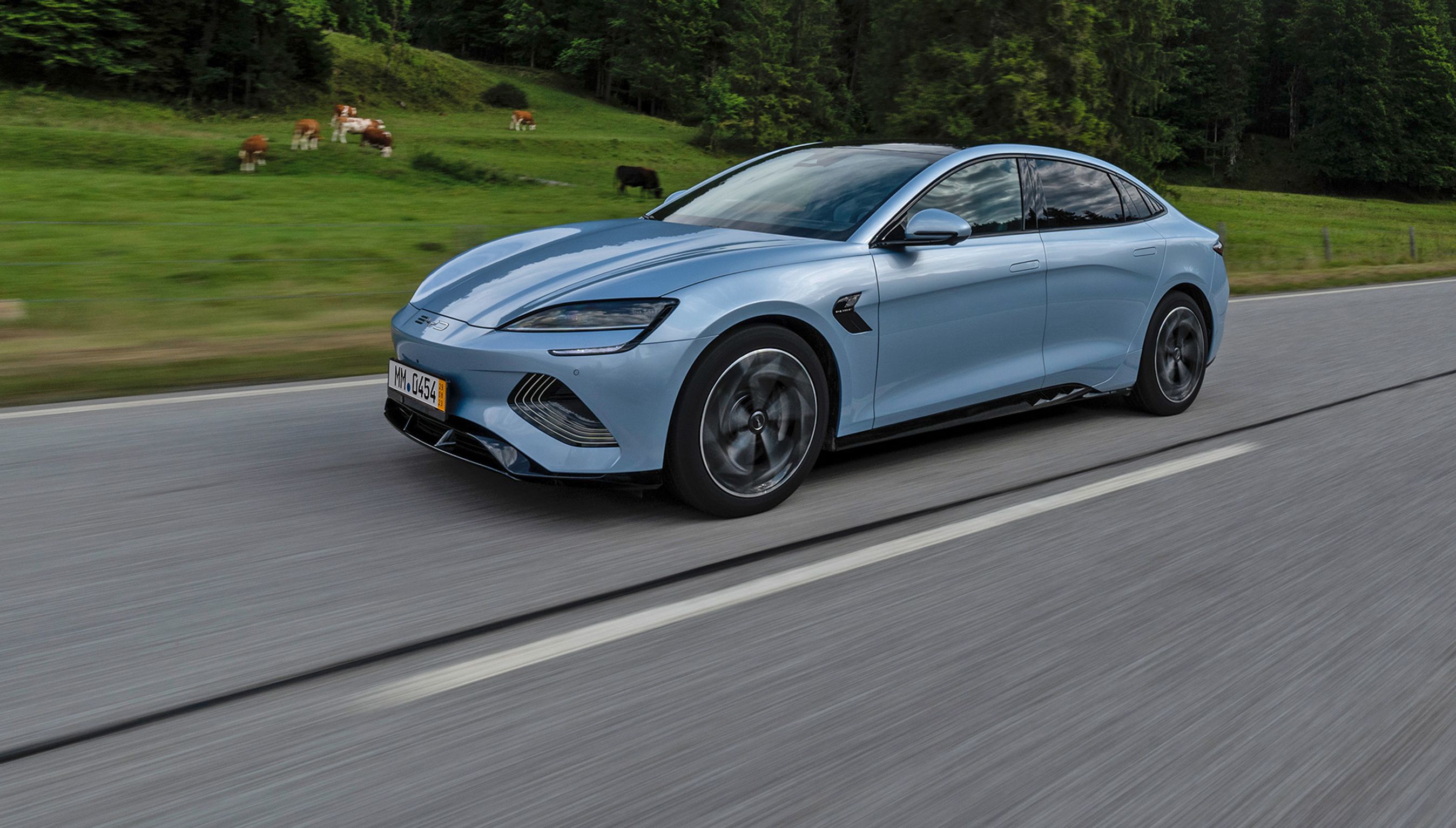 En lyseblå BYD Seal kører på en landevej med skove og bjerge i baggrunden