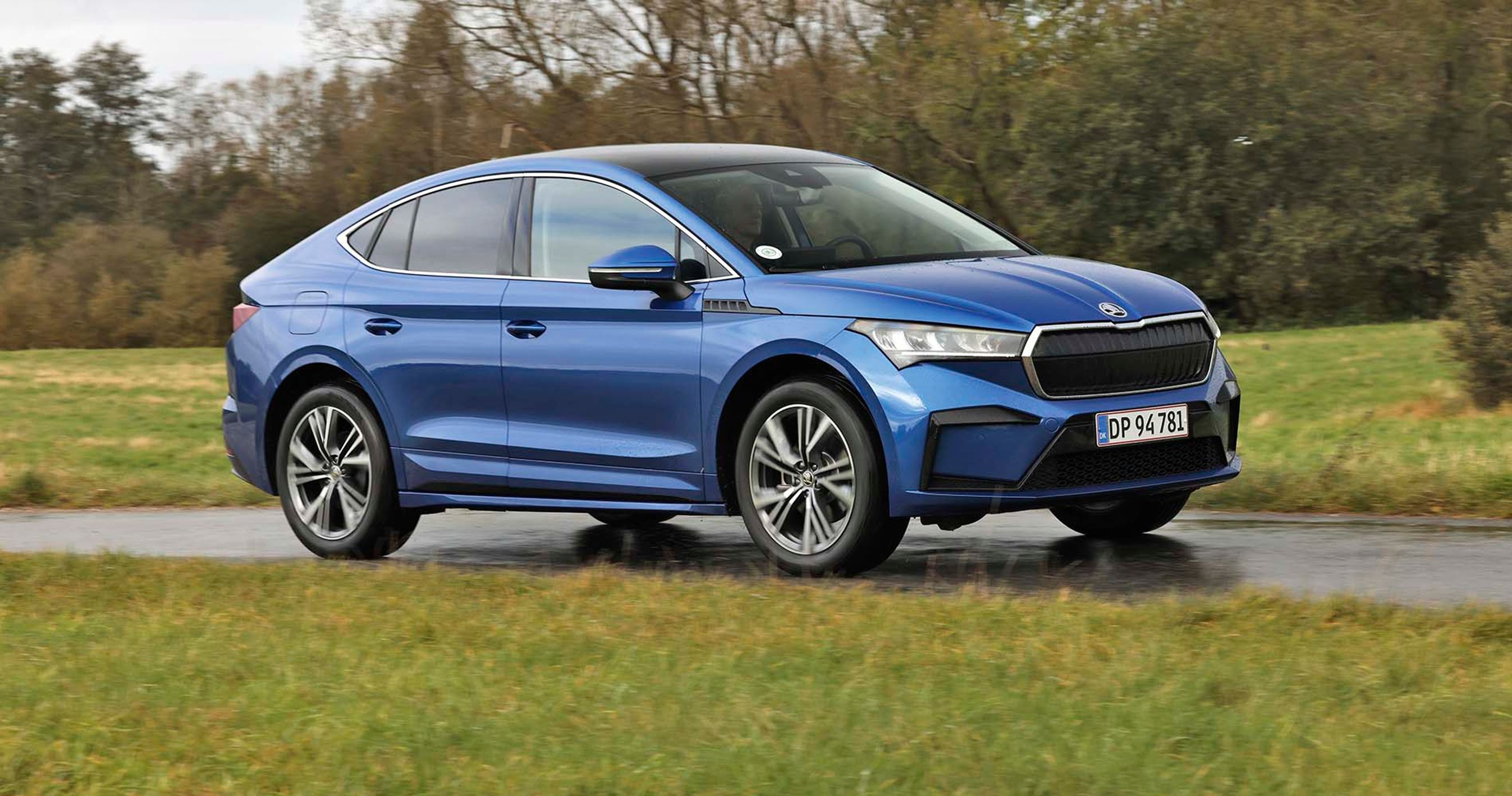 En blå Skoda Enyaq Coupé set fra siden, mens den kører på en landevej med græs i forgrunden