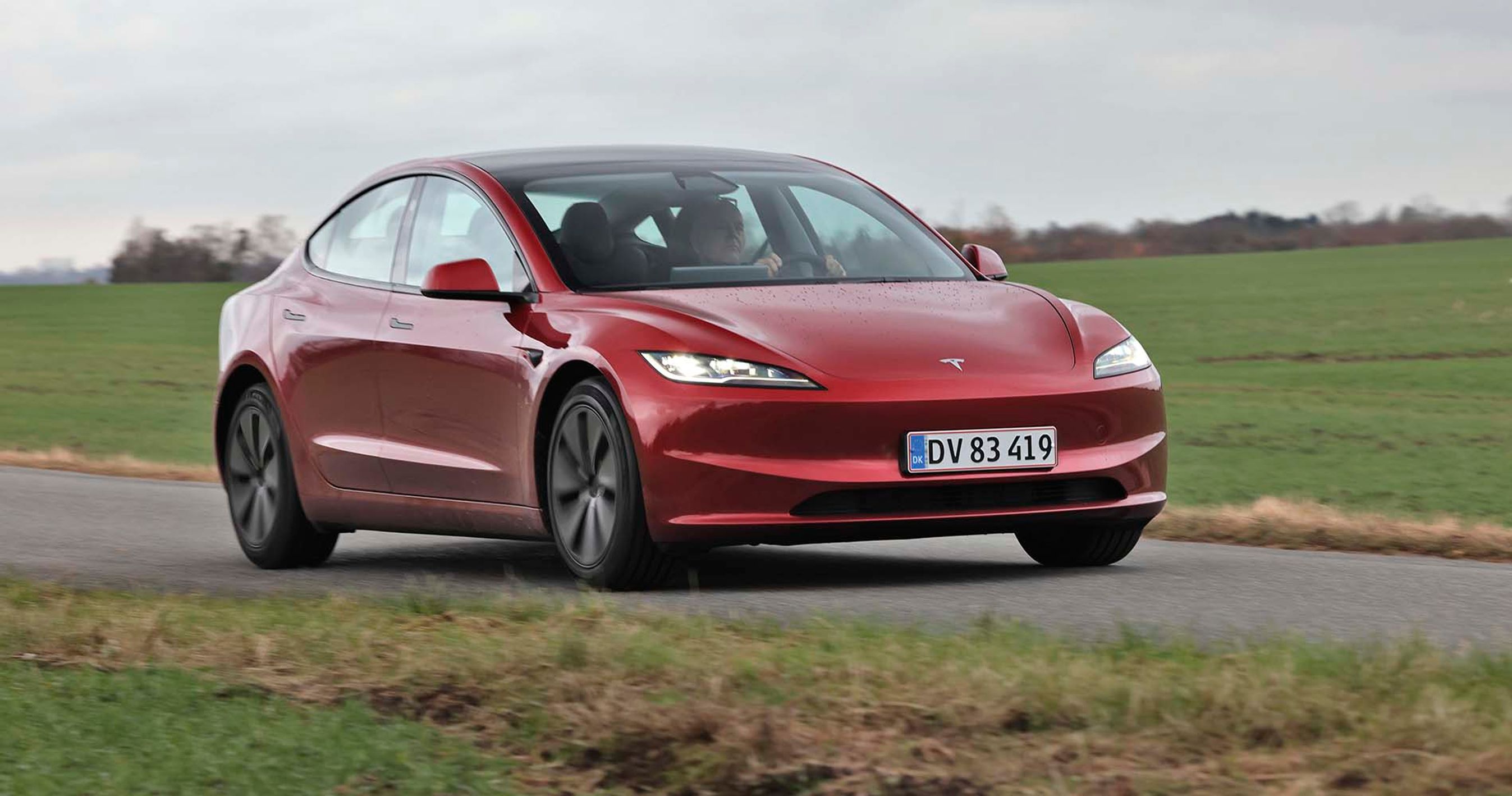 En rød Tesla Model 3 kører på en landevej med græs i baggrunden.