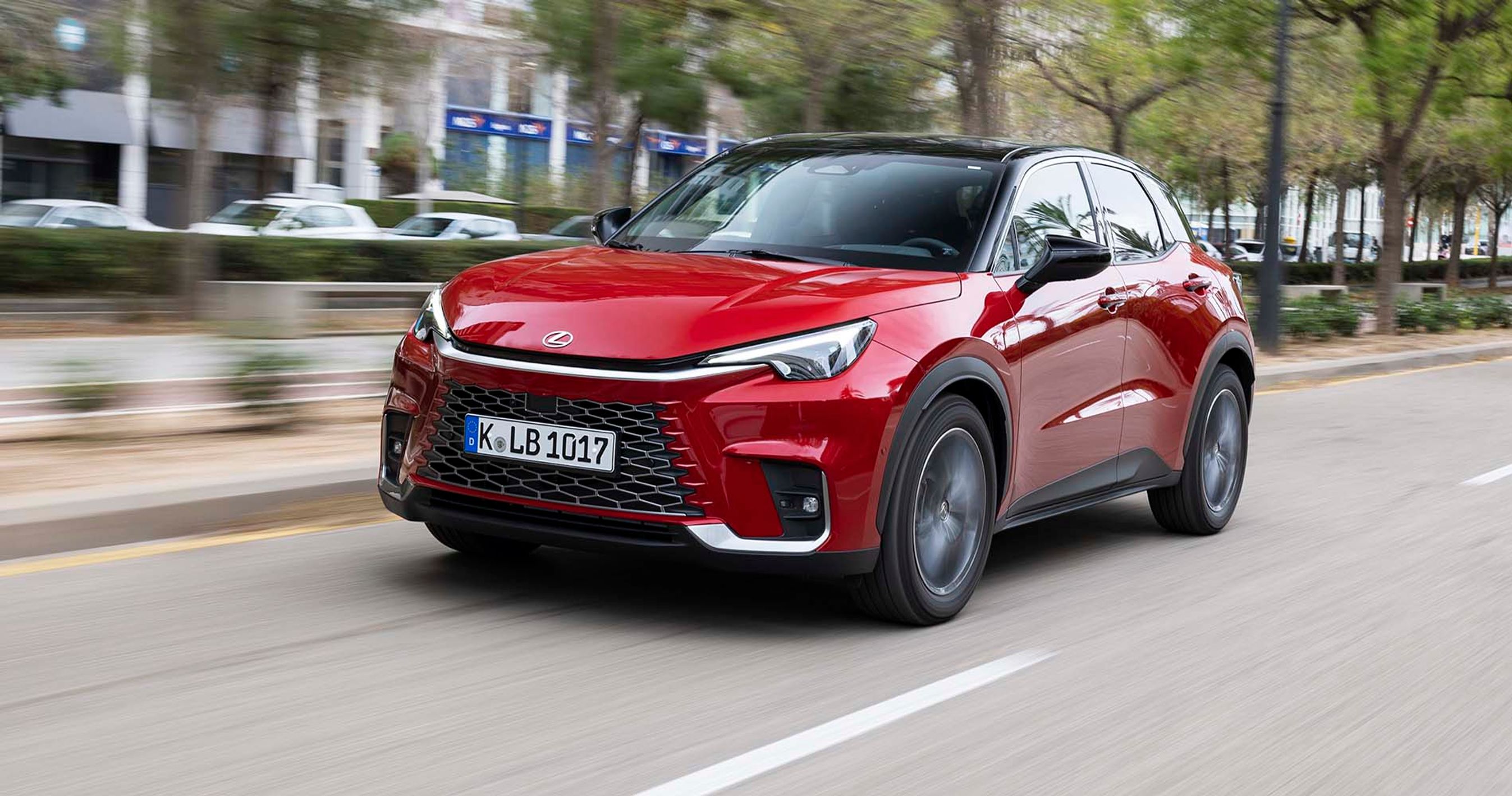 En rød Lecus LBX kører i en by med træer og bygninger i baggrunden