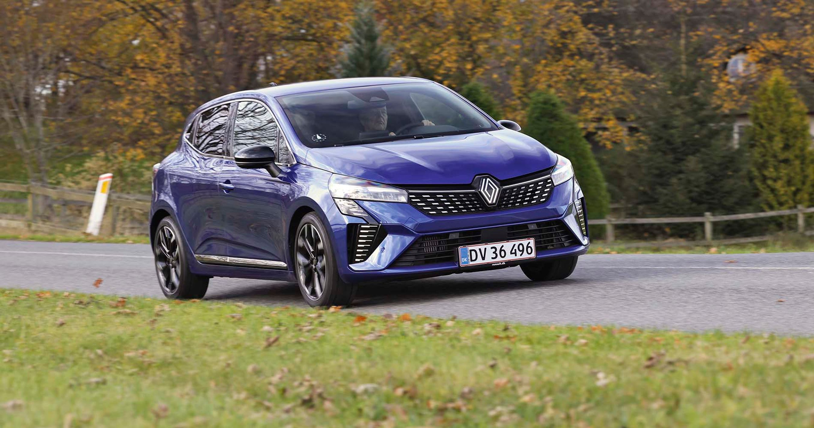 En blå Renault Clio kører på en landevej med græs i forgrunden og træer i baggrunden