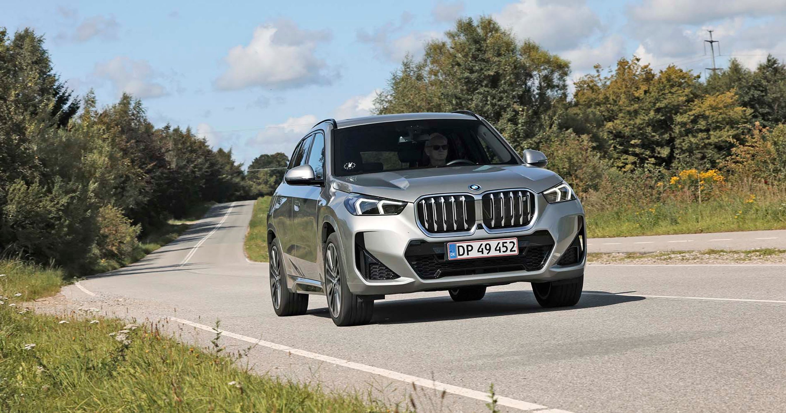 Elbilen BMW iX1 kører i et sving på en landevej med træer i baggrunden.