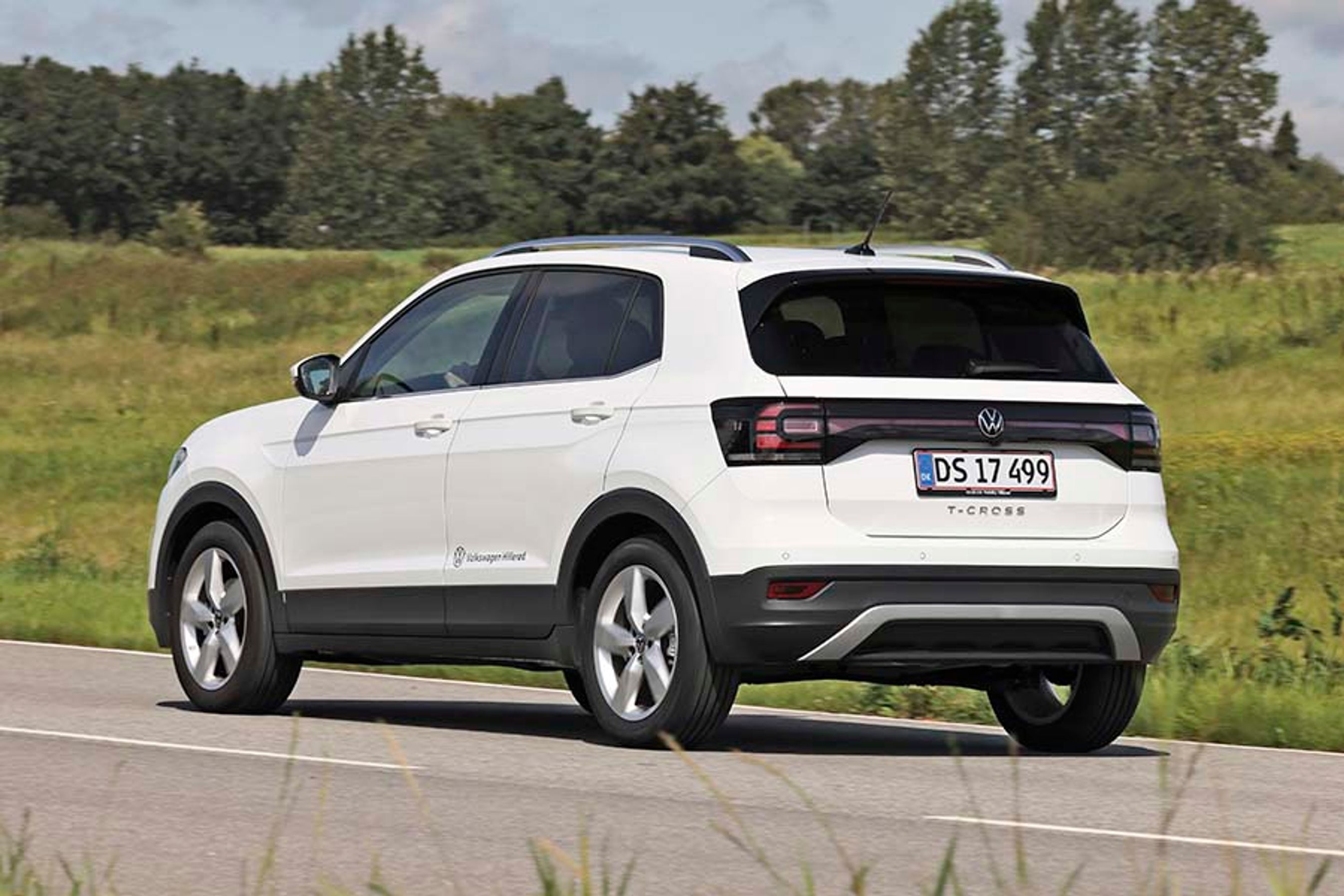 En hvid VW T-Cross set bagfra, mens den kører på en landevej med græs i baggrunden.