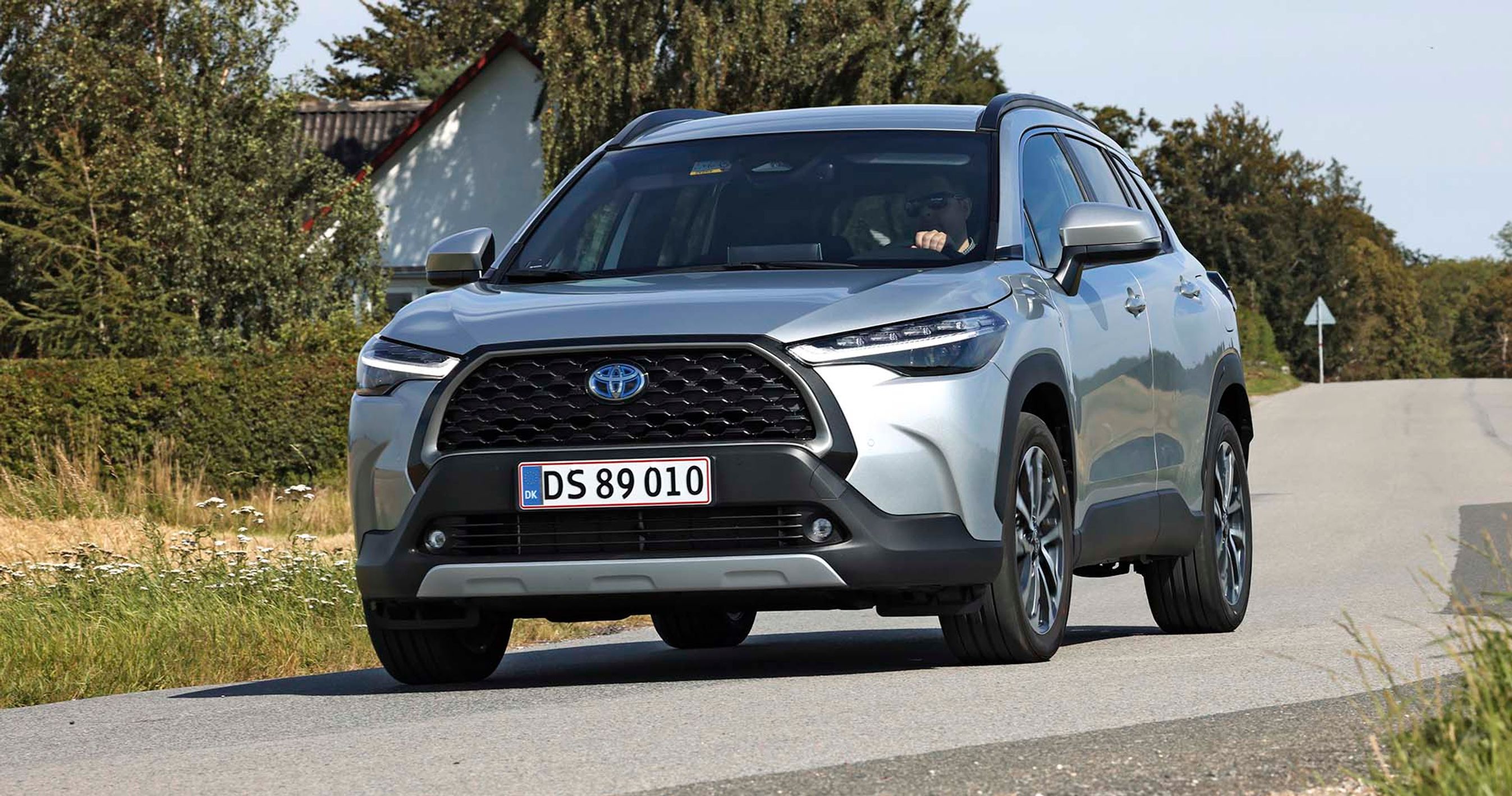 Hybridbilen Toyota Corolla Cross kører i et sving med et hvidt hus og træer i baggrunden.