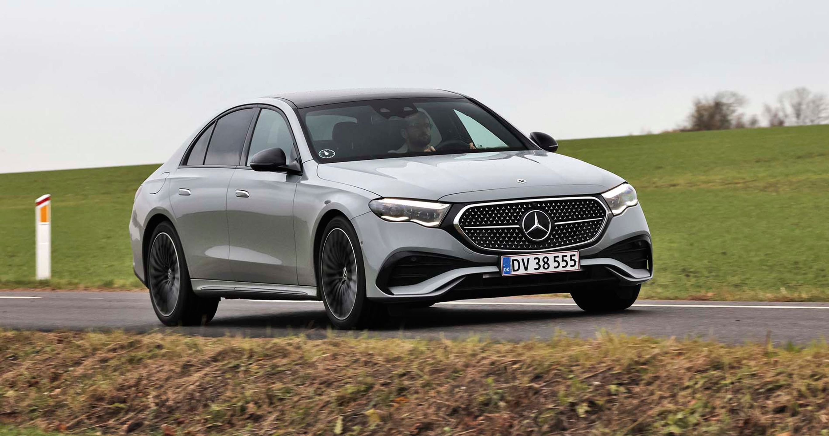 En sølvgrå Mercedes E-klasse set forfra, mens den kører på en vej med græs i baggrunden