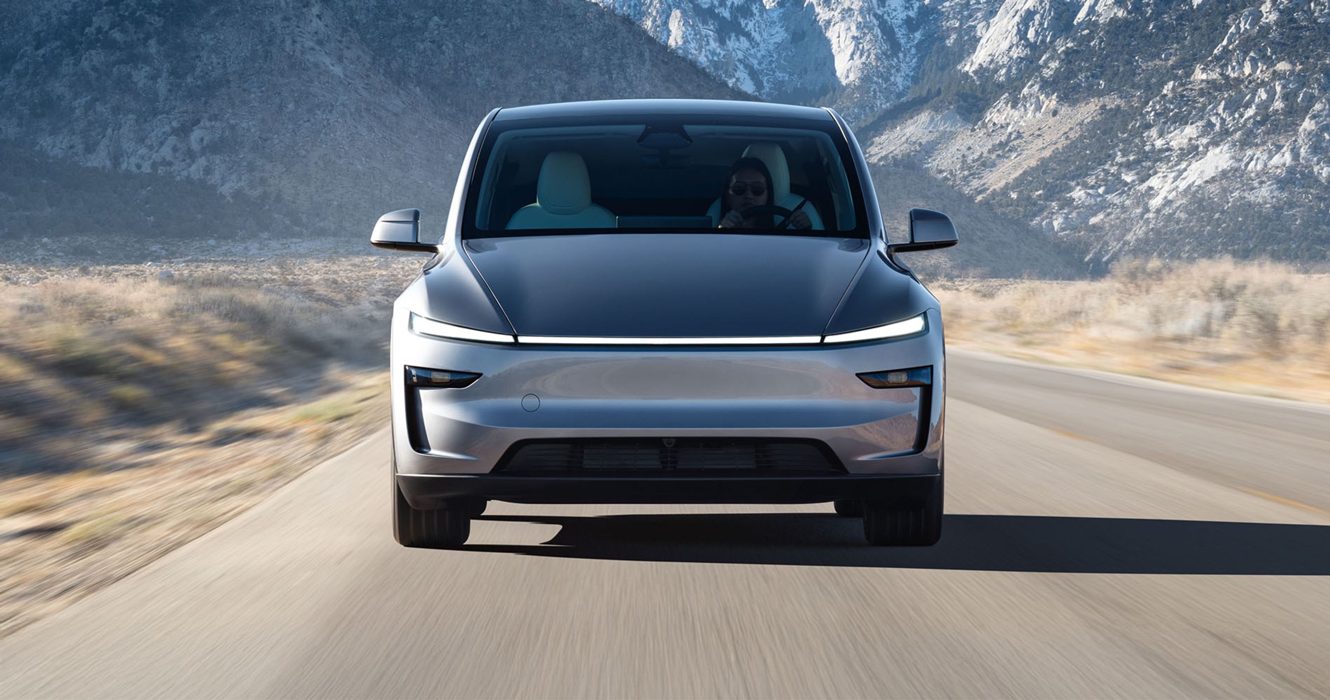 En grå faceliftet Tesla Model Y kører på vej med bjerge i baggrunden. 