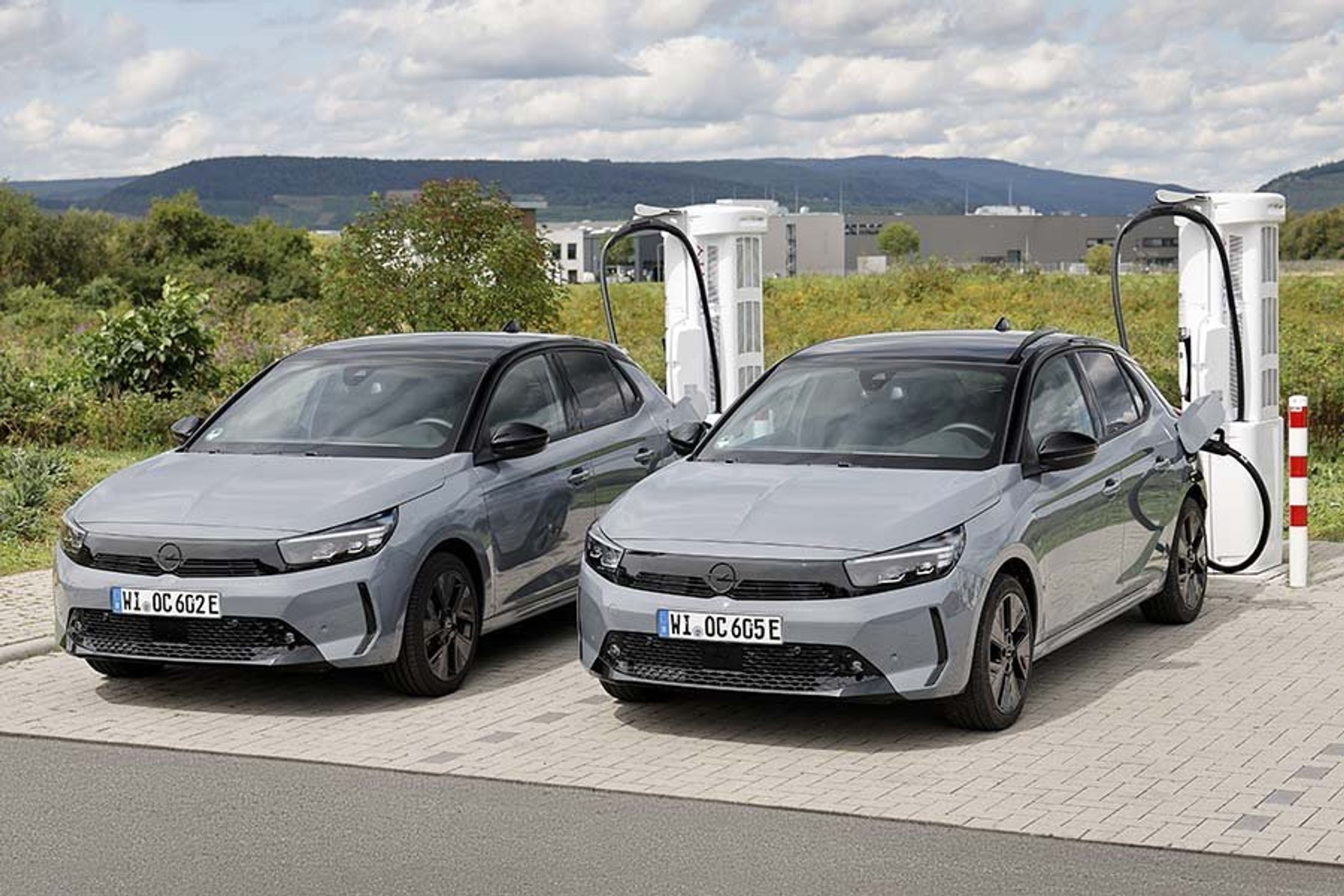 To Opel Corsa holder og lader ved en lynlader med bjerglandskab i baggrunden.