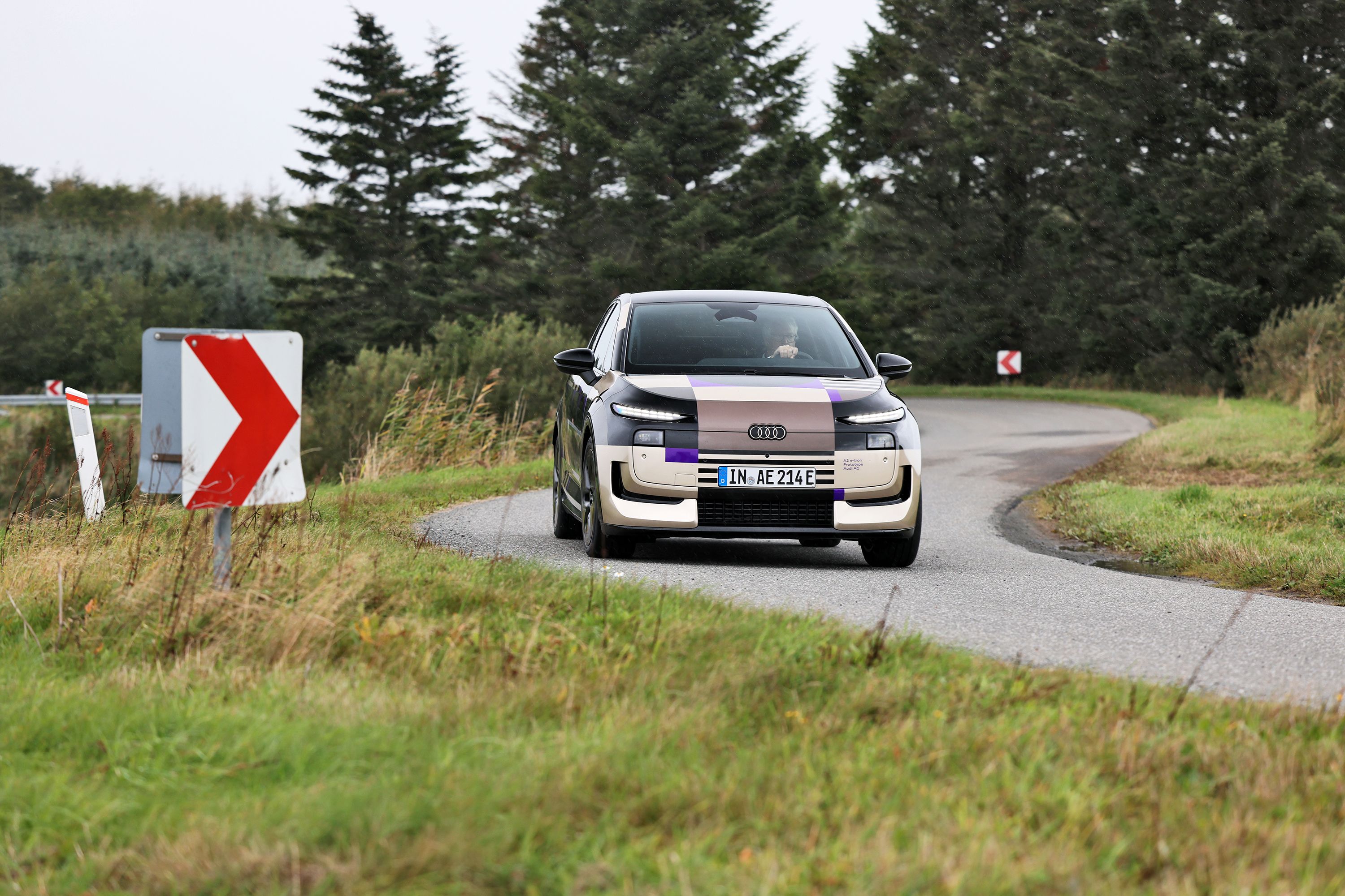 Audi A2 kører på dansk landevej med træer i baggrunden og skilte med pile på. 
