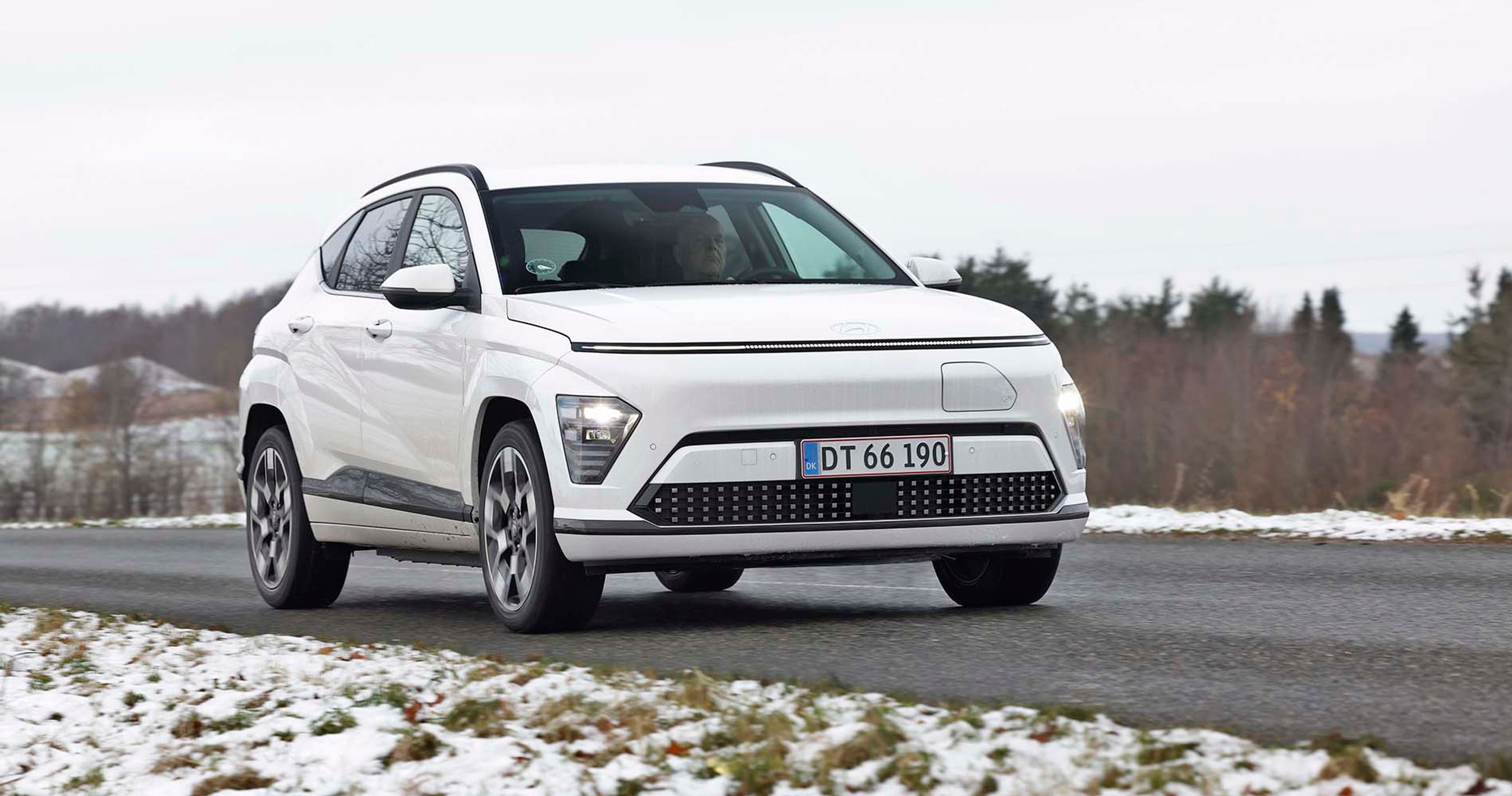 En hvid Hyundai Kona elbil kører på en landevej på en gråvejrsdag