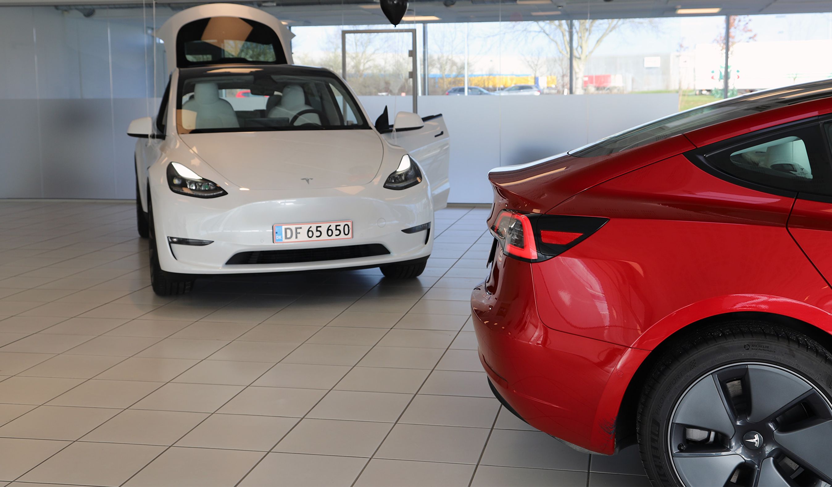 Hvid bil forfra, rød bil i forgrunden.