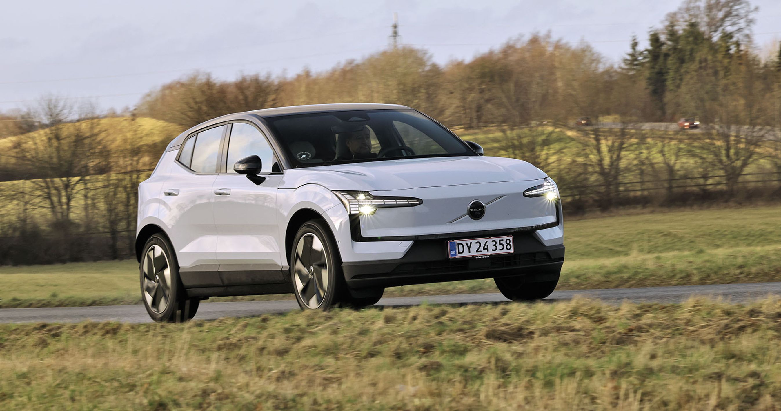 En hvid Volvo EX30 kører på en landevej med græs på begge sider