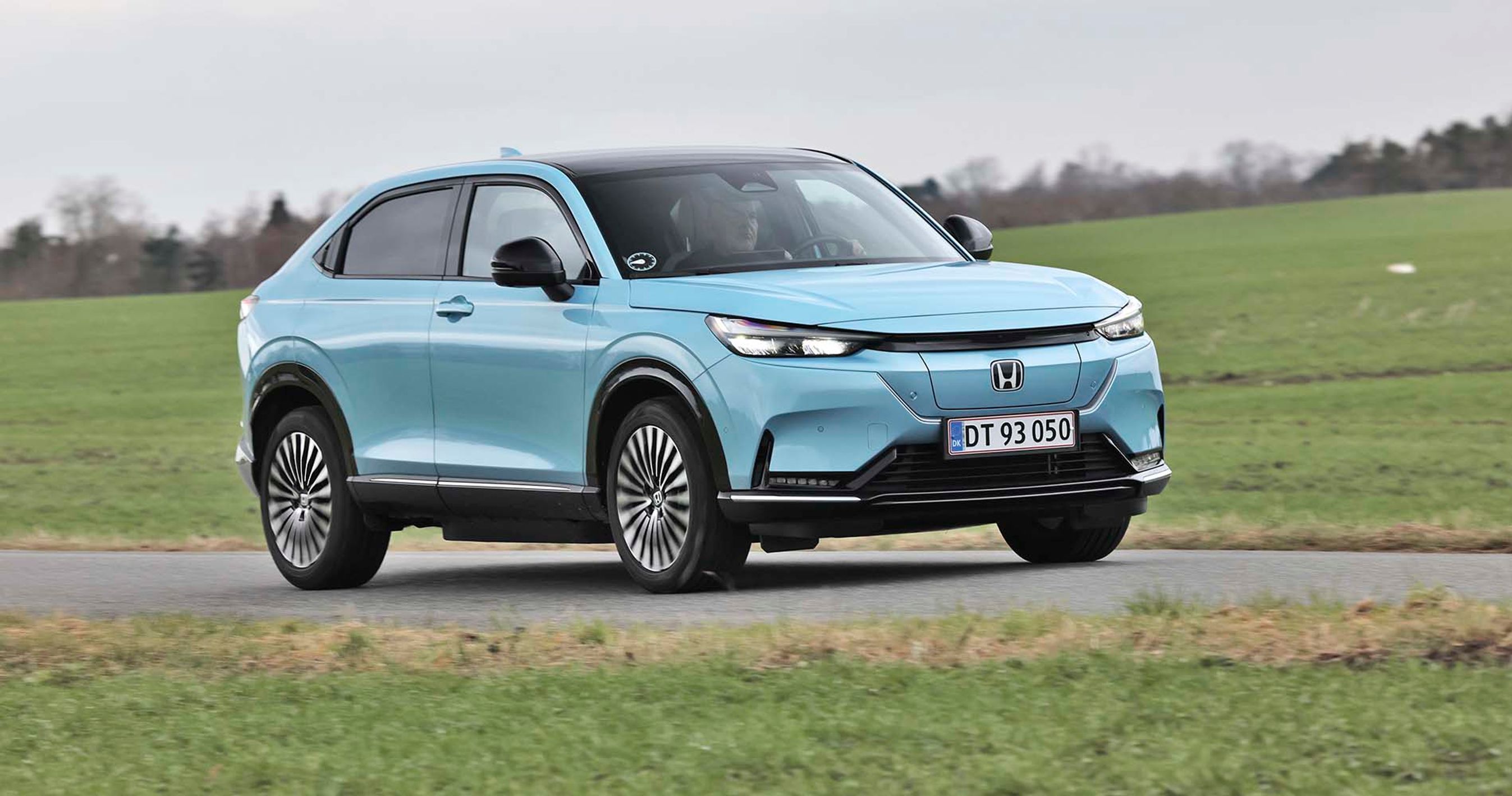En isblå Honda e:NY1 elbil kører på en landevej med græs rundt om