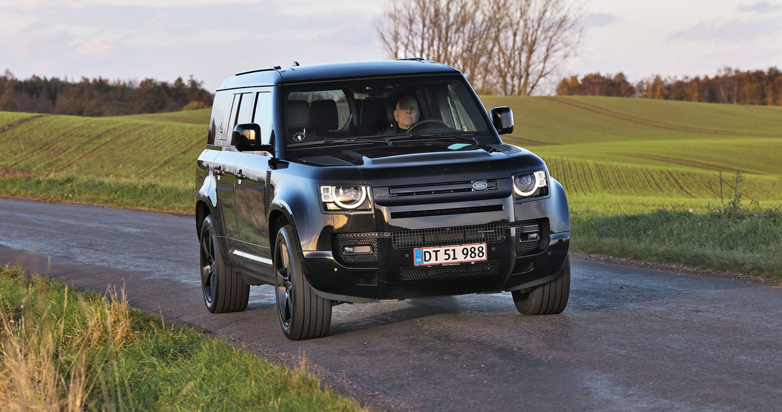 Land Rover Defender kører på en landevej med græs i baggrunden.