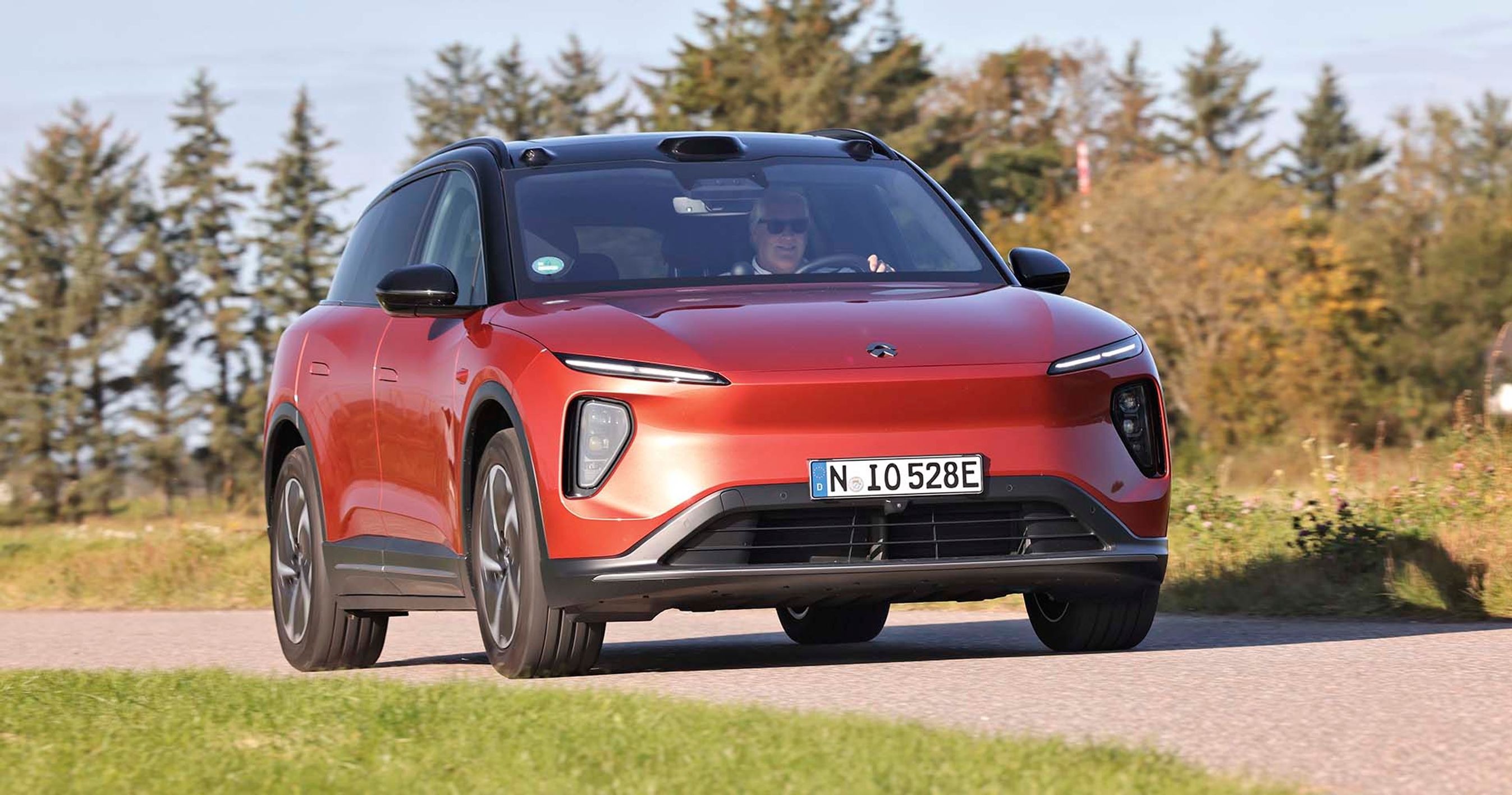 En rød Nio EL6 elbil kører på en landevej med træer i baggrunden.