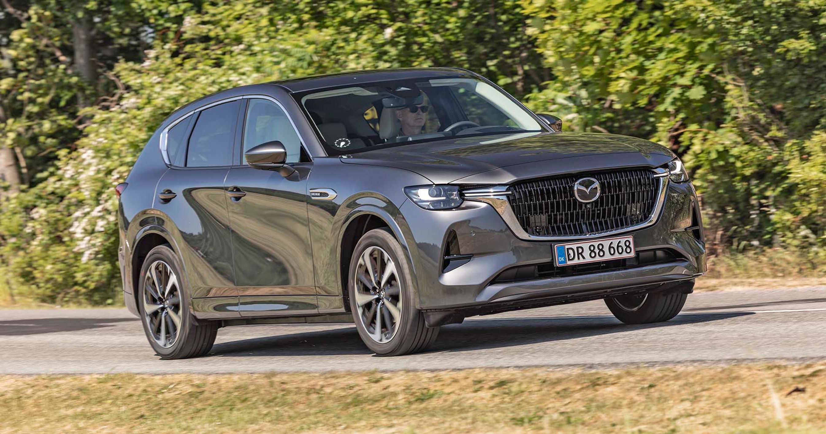 En Mazda CX-60 med stor dieselmotor kører på en landevej med træer i baggrunden