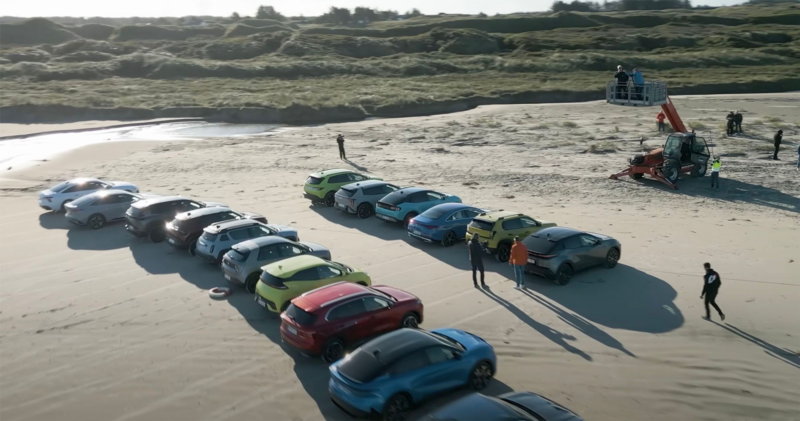 Mange biler står på stranden og der holder en kran med to personer i højre side. 