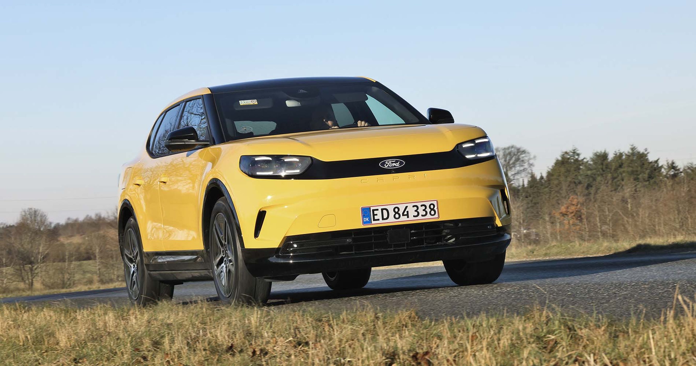 Gul Ford Capri set forfra, mens den kører på en landevej med græs i forgrunden og træer i baggrunden