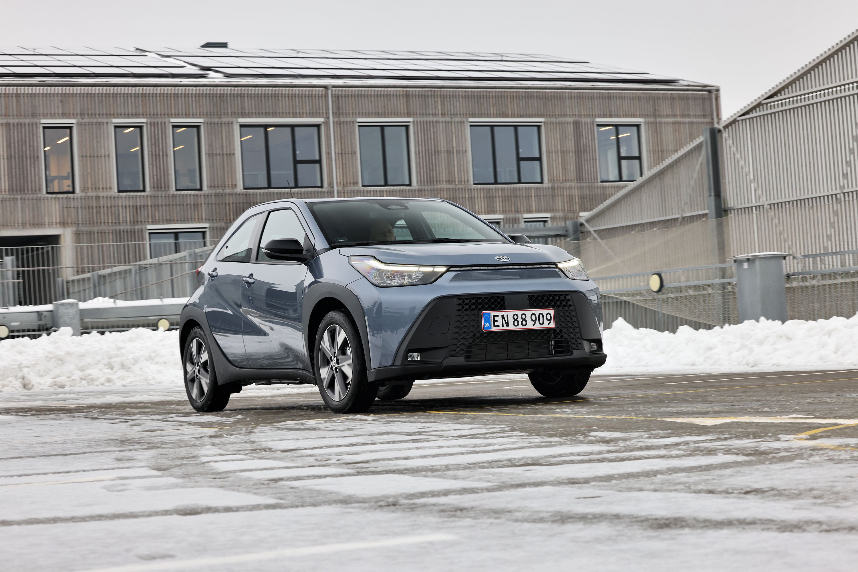 Toyota Aygo X set forfra, mens den holder foran bygning med trælister, og der er sne i baggrunden