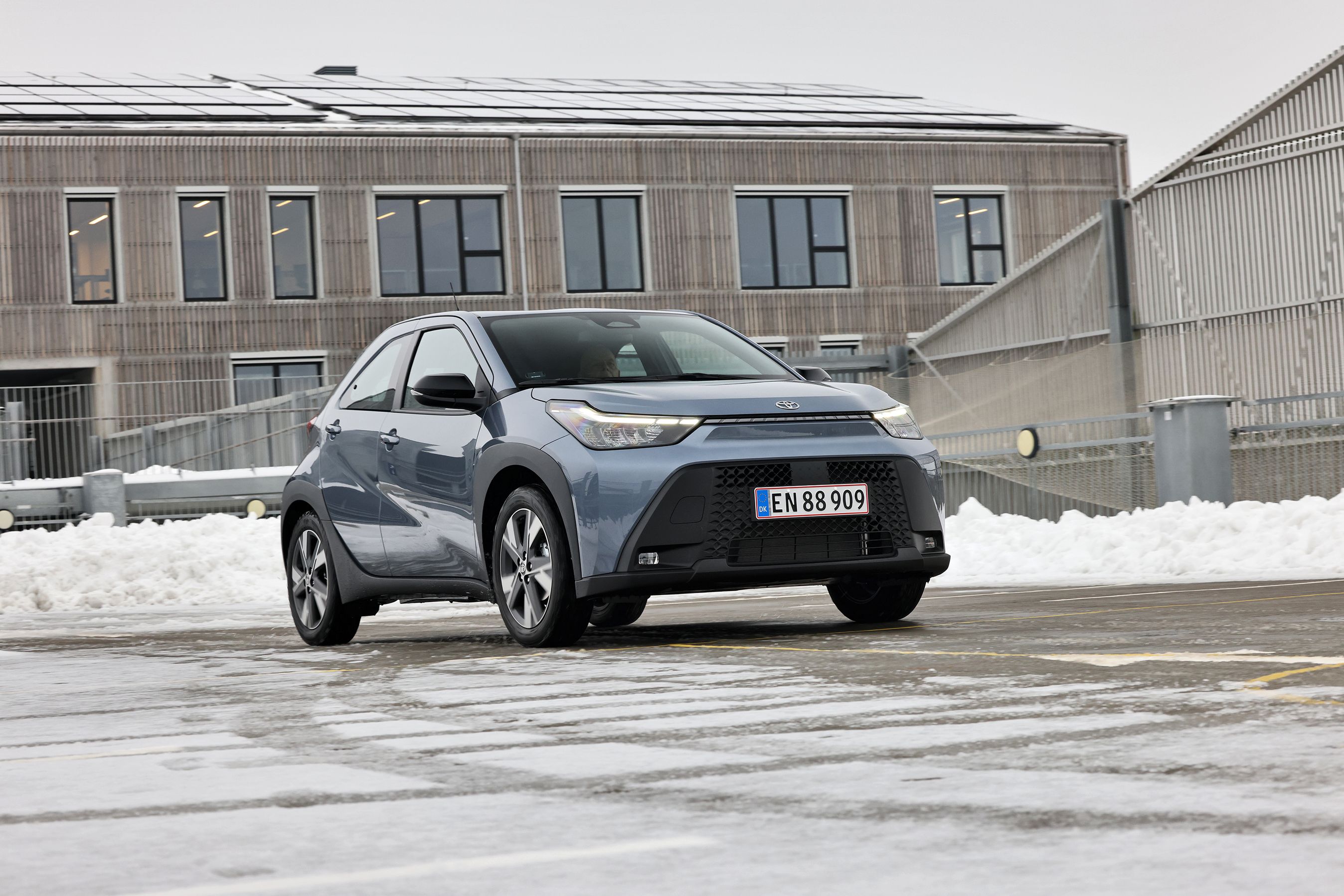 Toyota Aygo X set forfra, mens den holder foran bygning med trælister, og der er sne i baggrunden