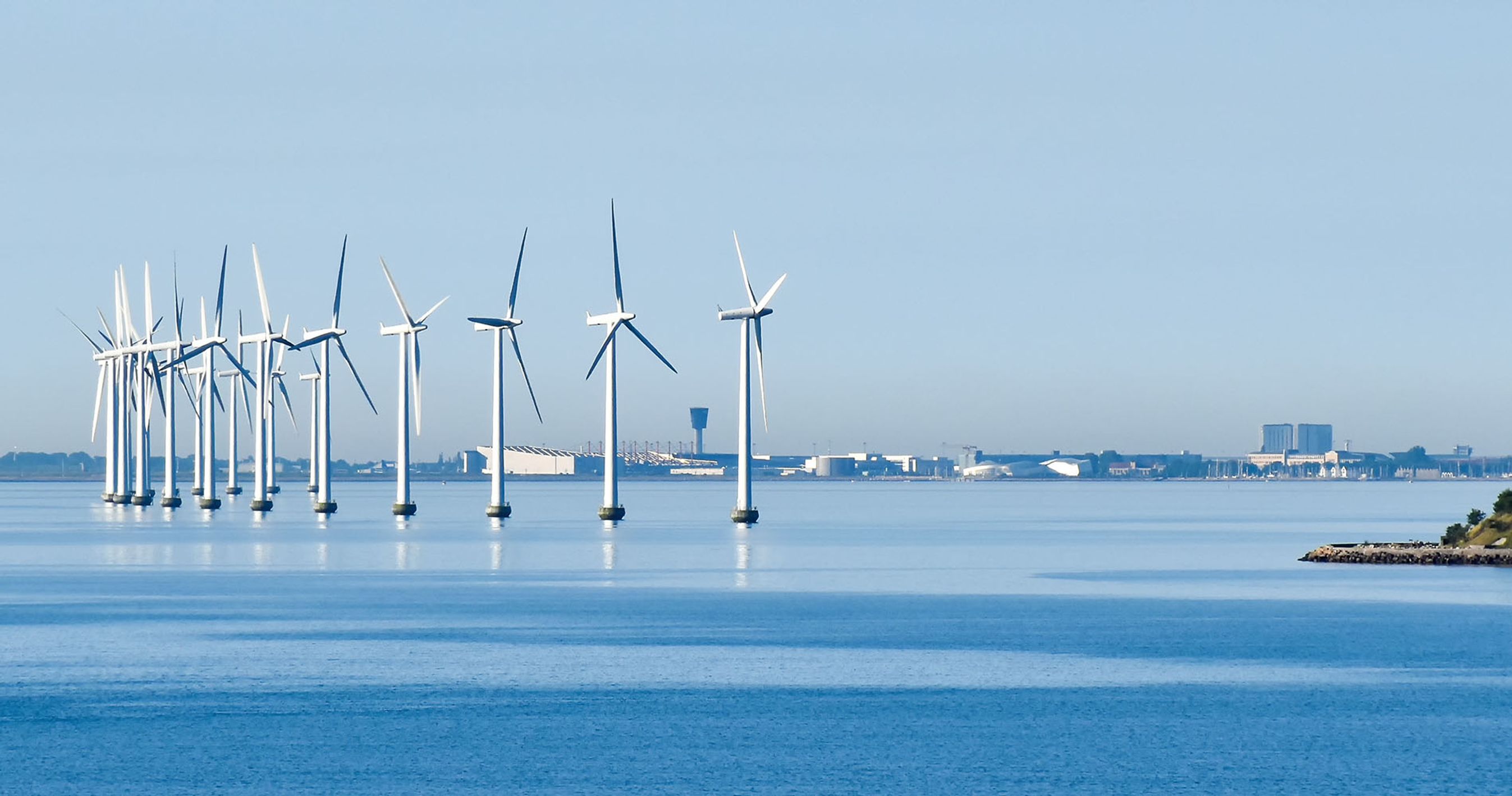 Vindmøller står på havet med kystlinjen i baggrunden.