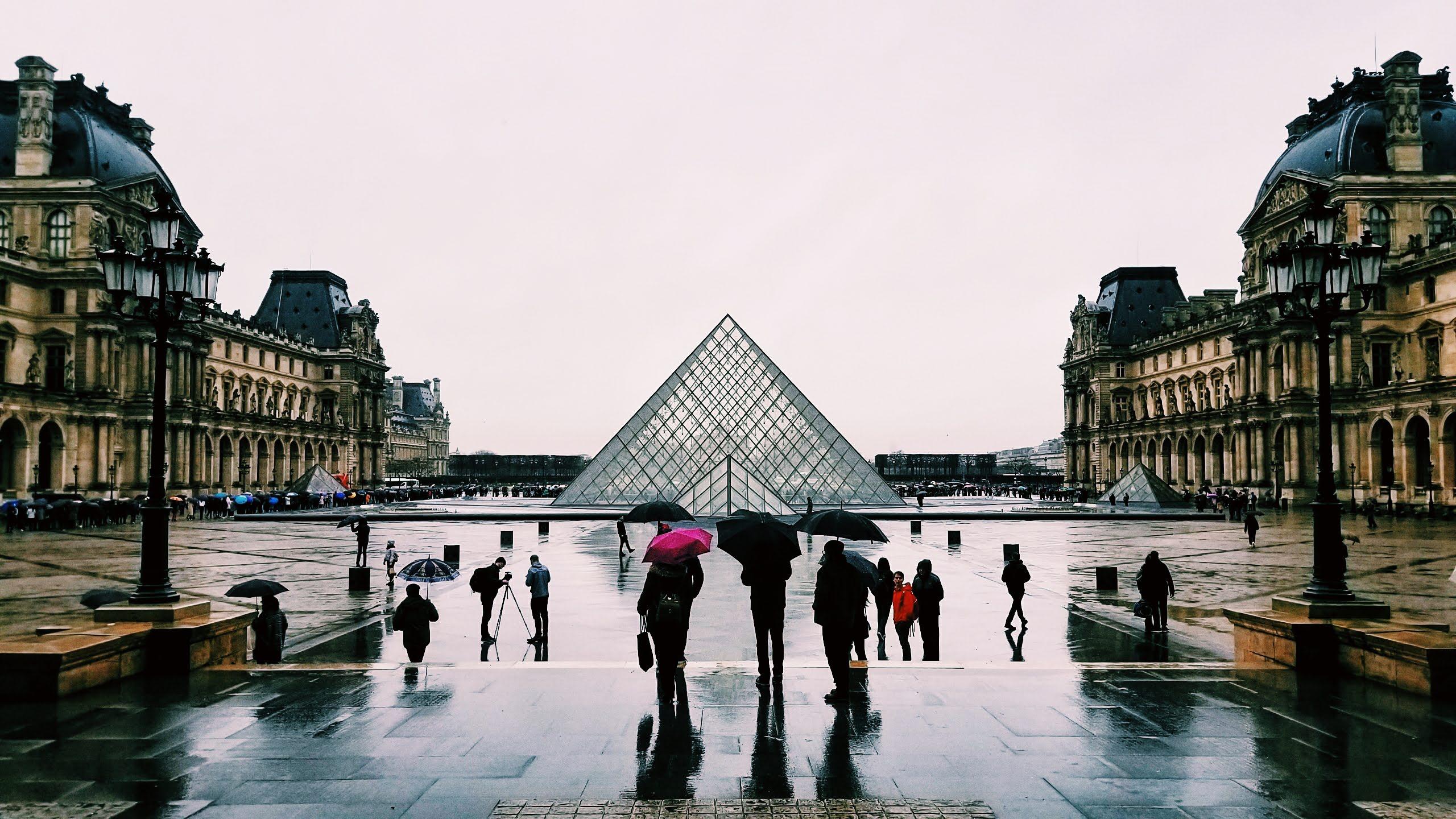 Tourists with umbrellas looking at the Pyramids of the Louvre on a rainy day.