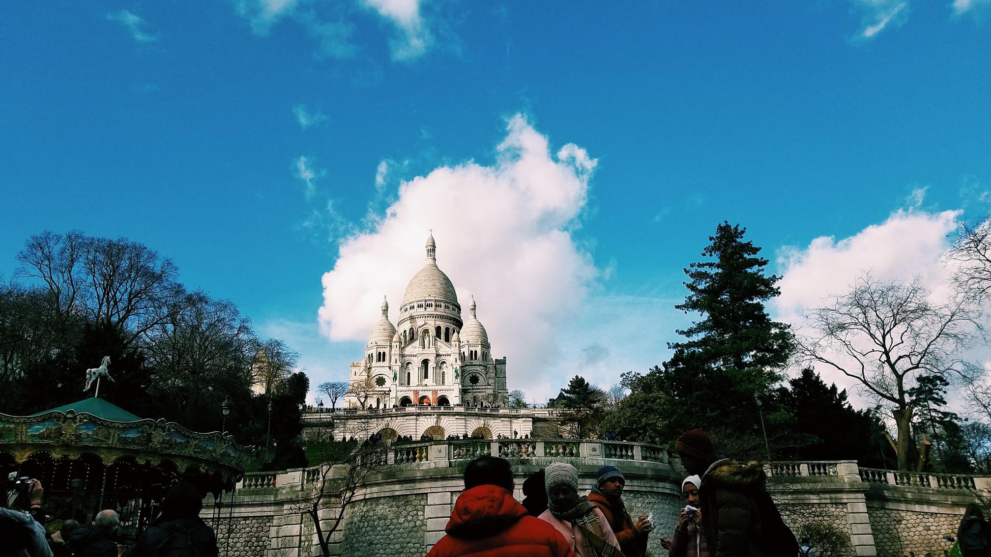 Sacre Coeur on a bright sunny day, sitting on the hill above some admiring tourists and the carousel to their left.