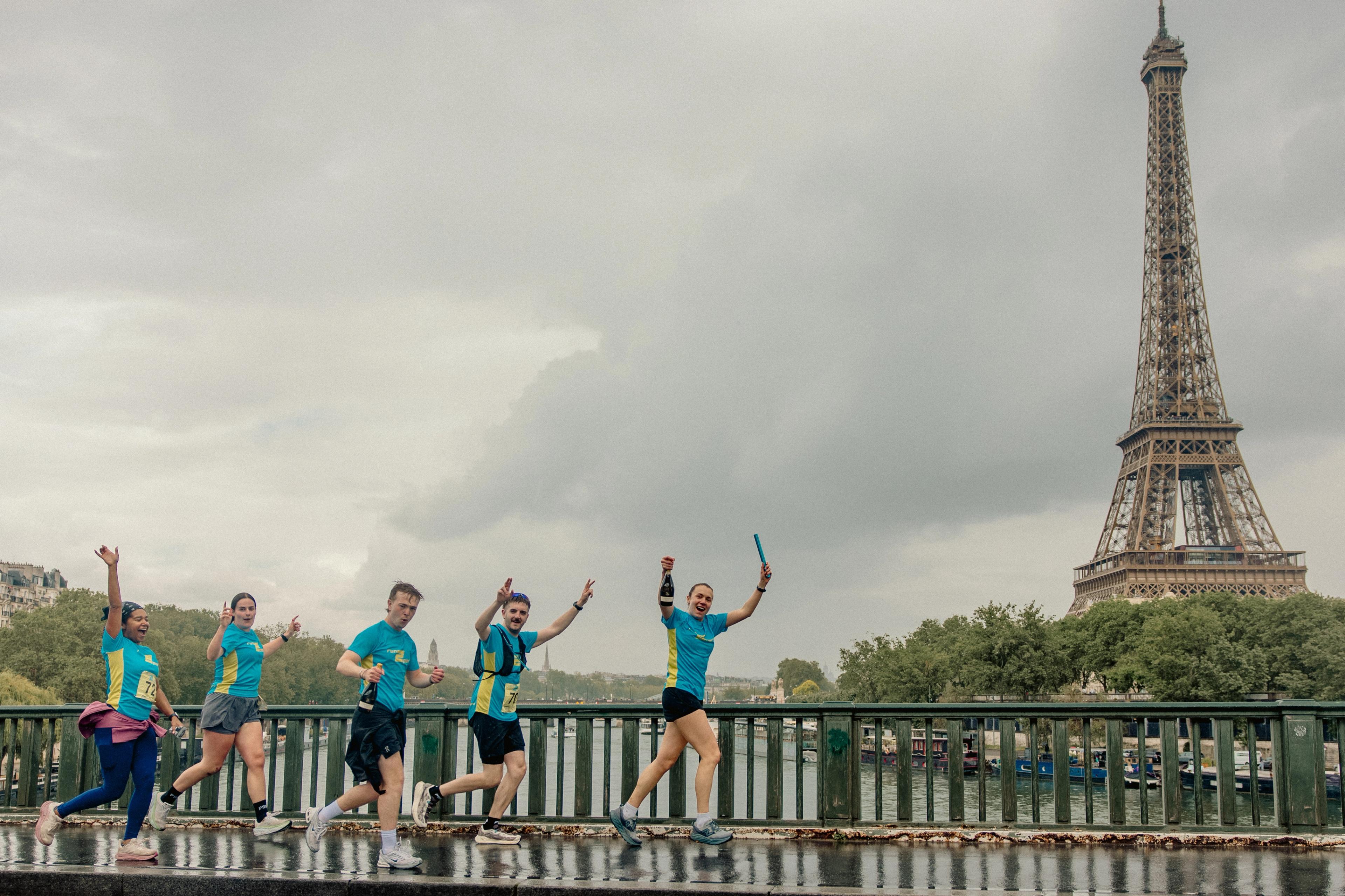 Five runners in blue and yellow shirts raise their arms on a wet bridge with the Eiffel Tower in the background.