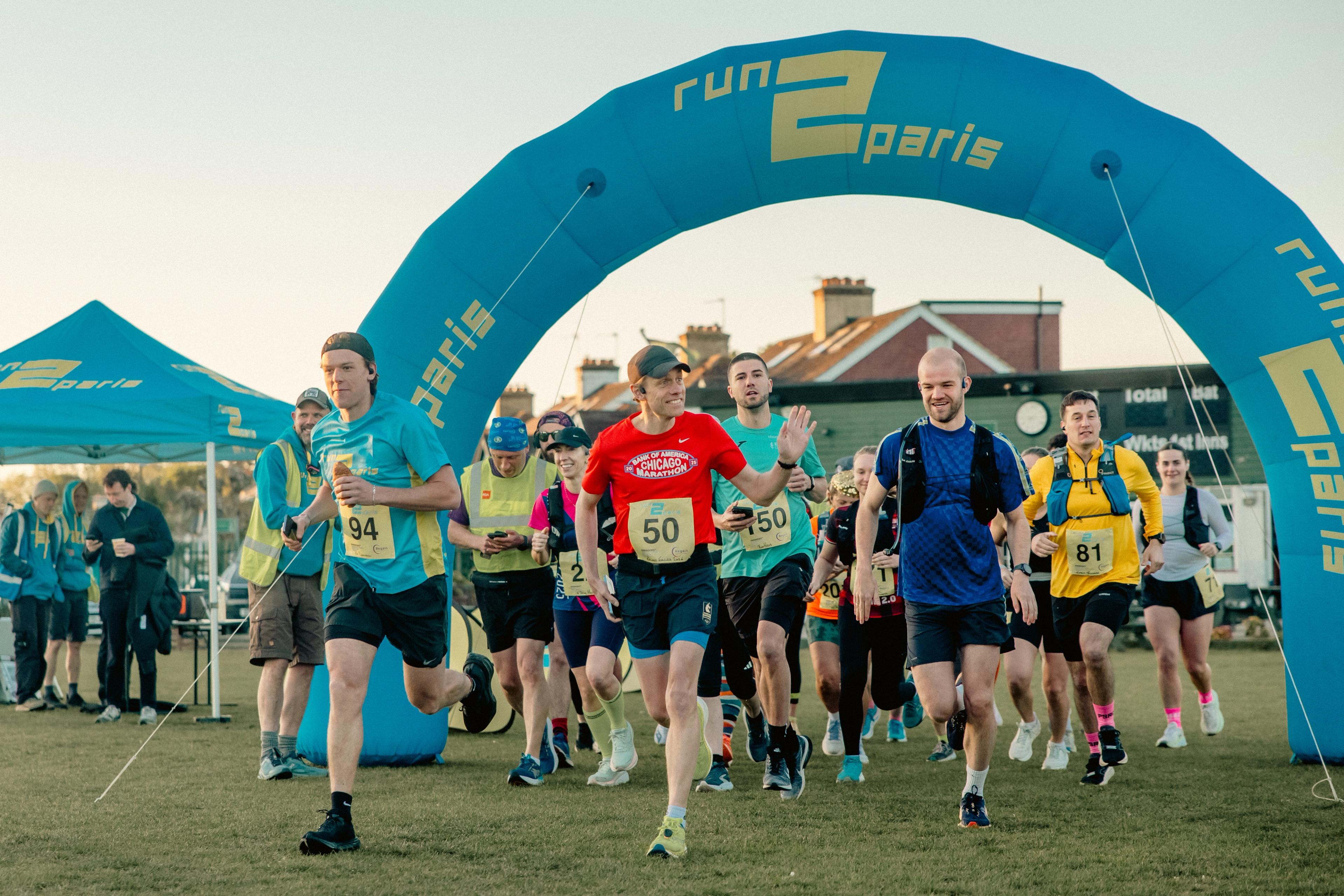 Runners starting a race under a blue "run 2 paris" inflatable arch on a grassy field.