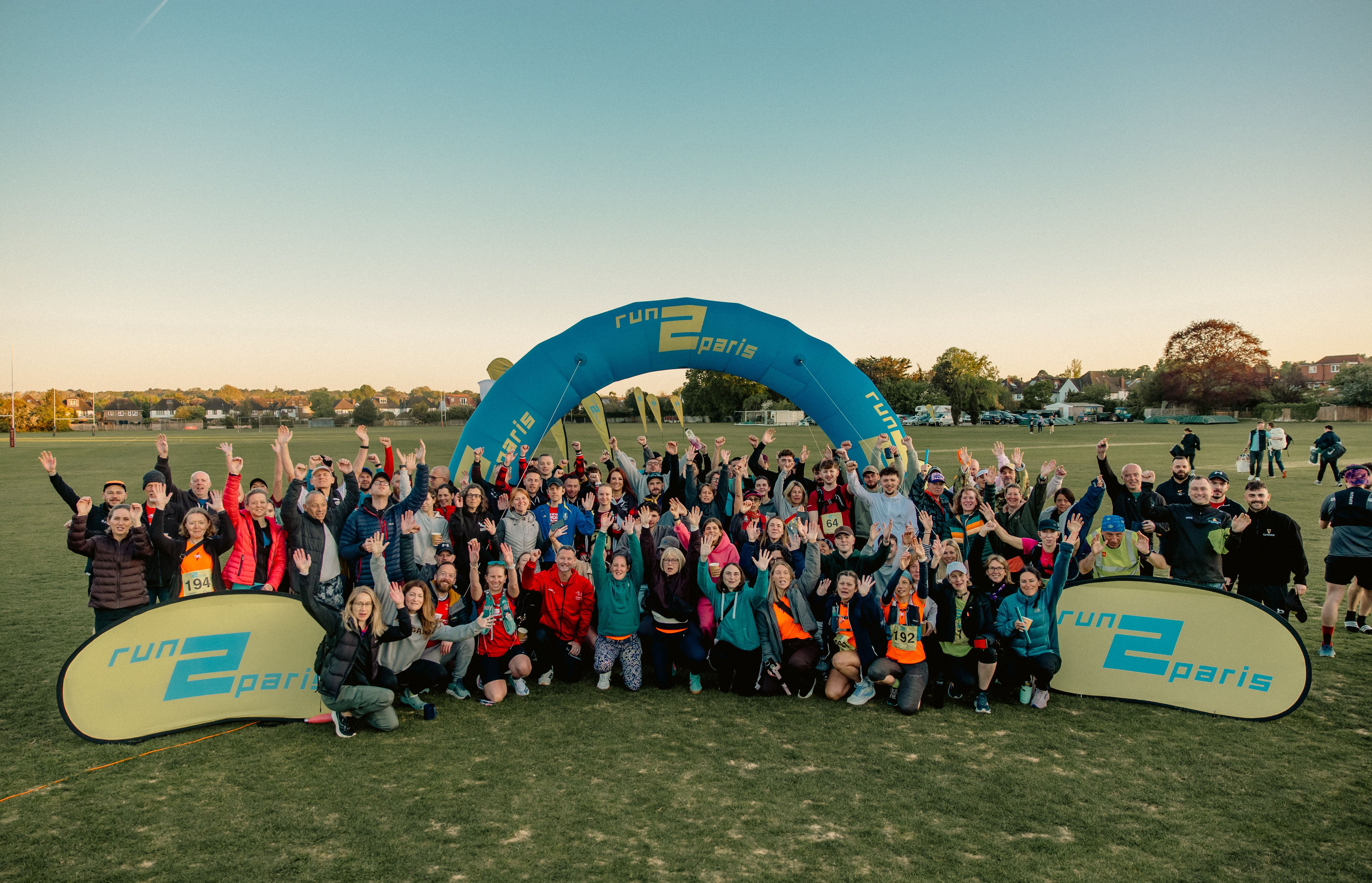 A large group of people poses happily on a grassy field in front of a blue "Run 2 Paris" inflatable arch.