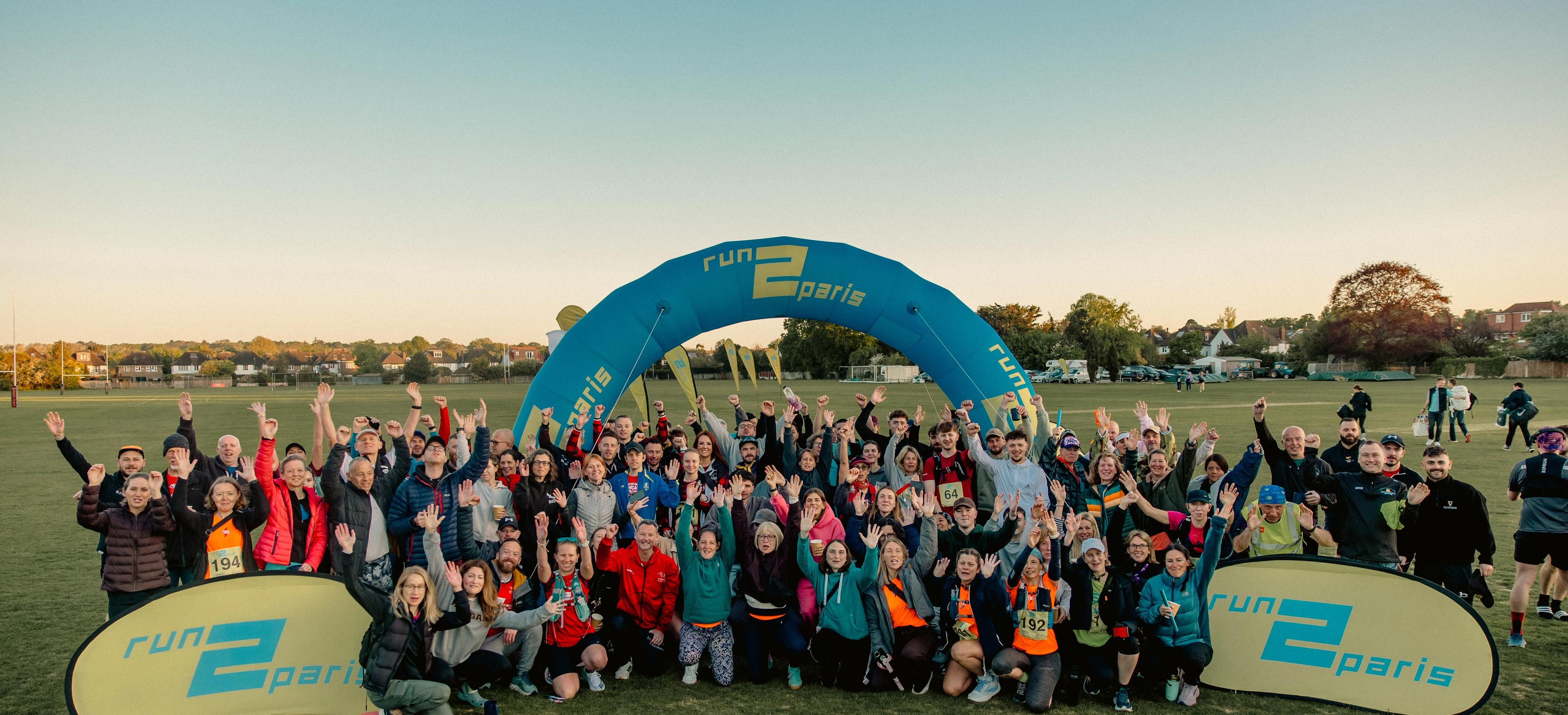A large group of people poses happily on a grassy field in front of a blue "Run 2 Paris" inflatable arch.