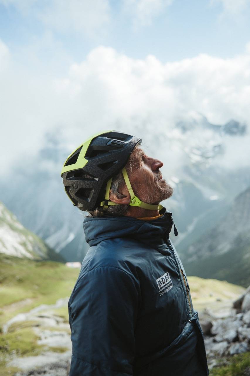 Person in a climbing helmet looking up, clouds and mountain slopes behind