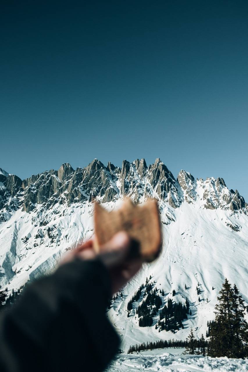 A hand holding up a piece of bread in front of snowy rock peaks and a deep blue sky