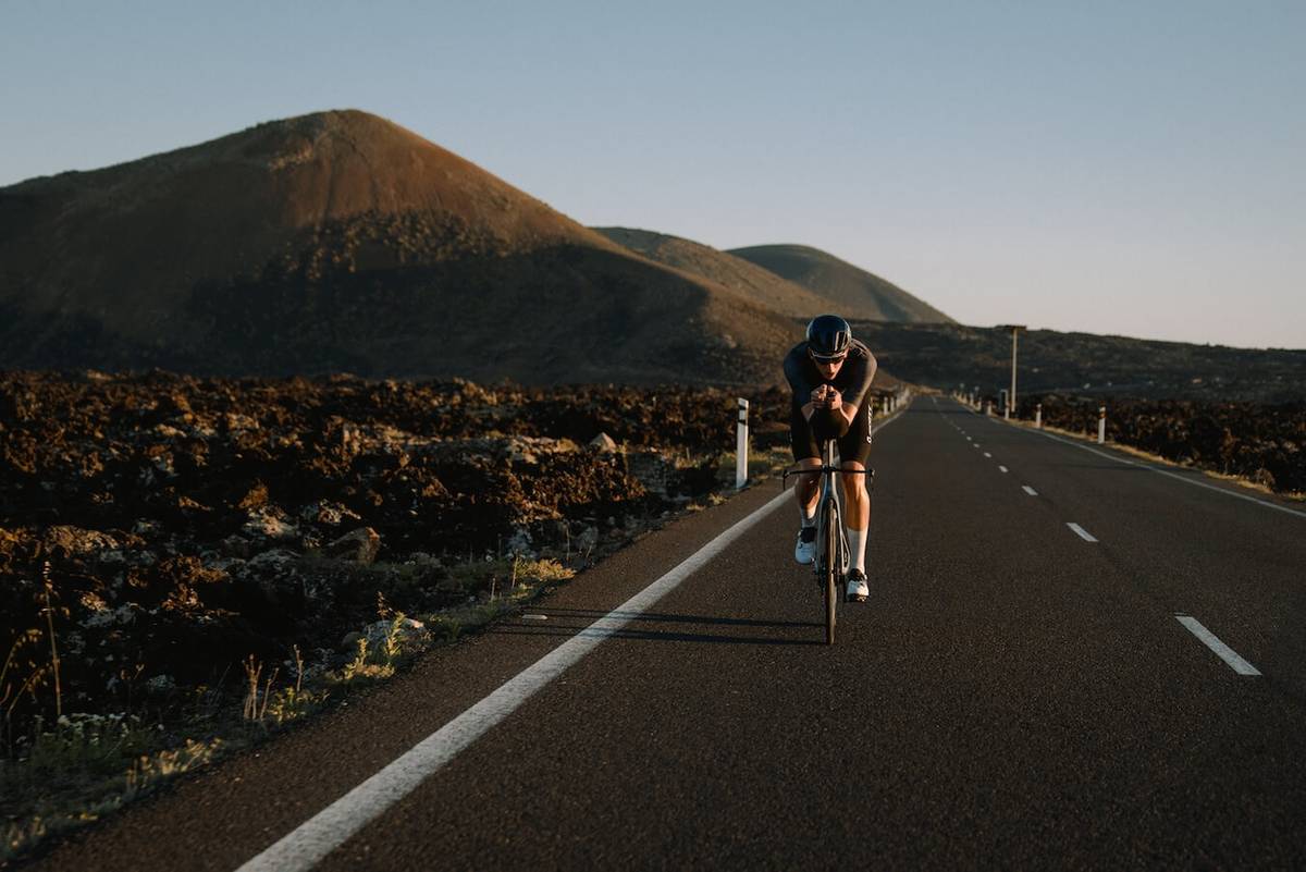 Cyclist coming head-on down a straight road through lava fields, a volcano behind him