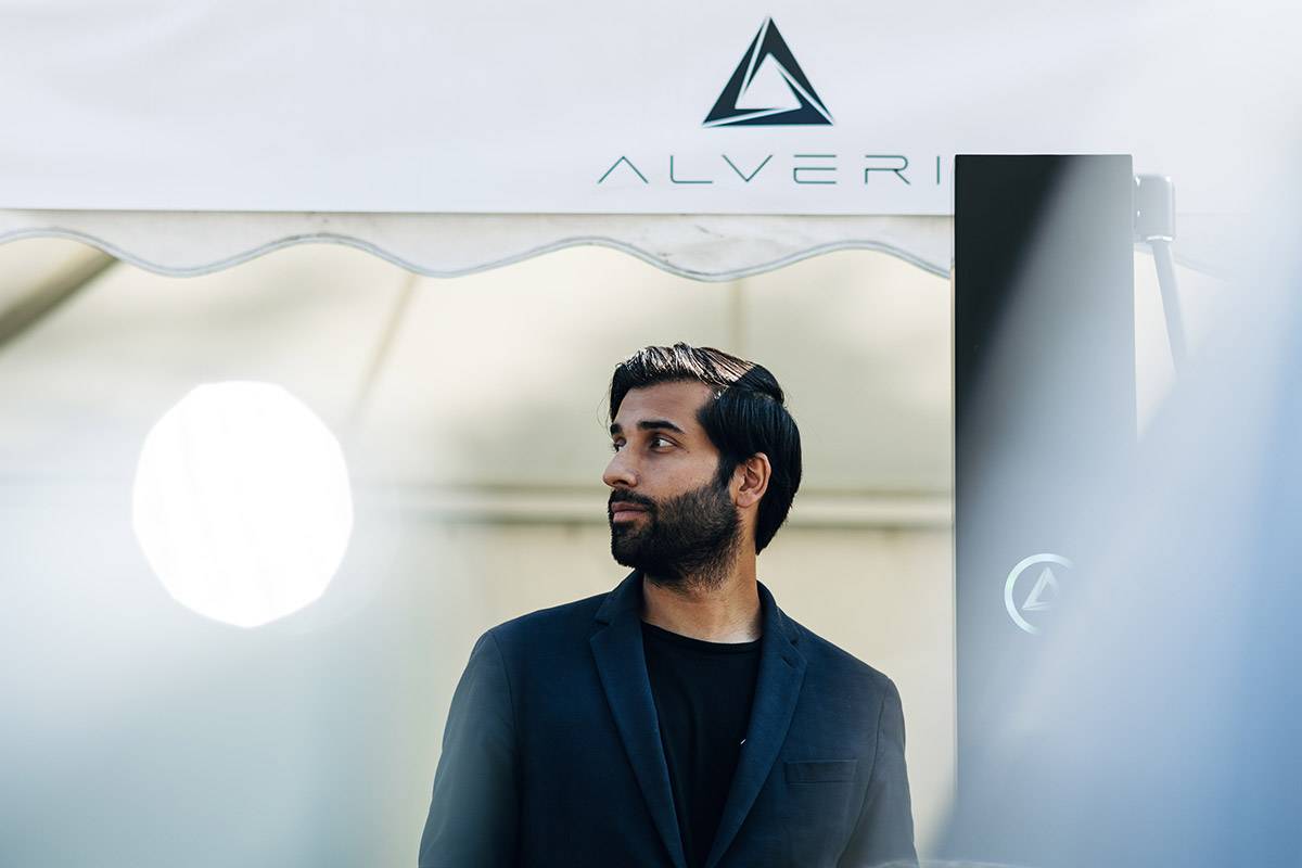 Man in a dark jacket looking to the side, the Alveri logo behind him