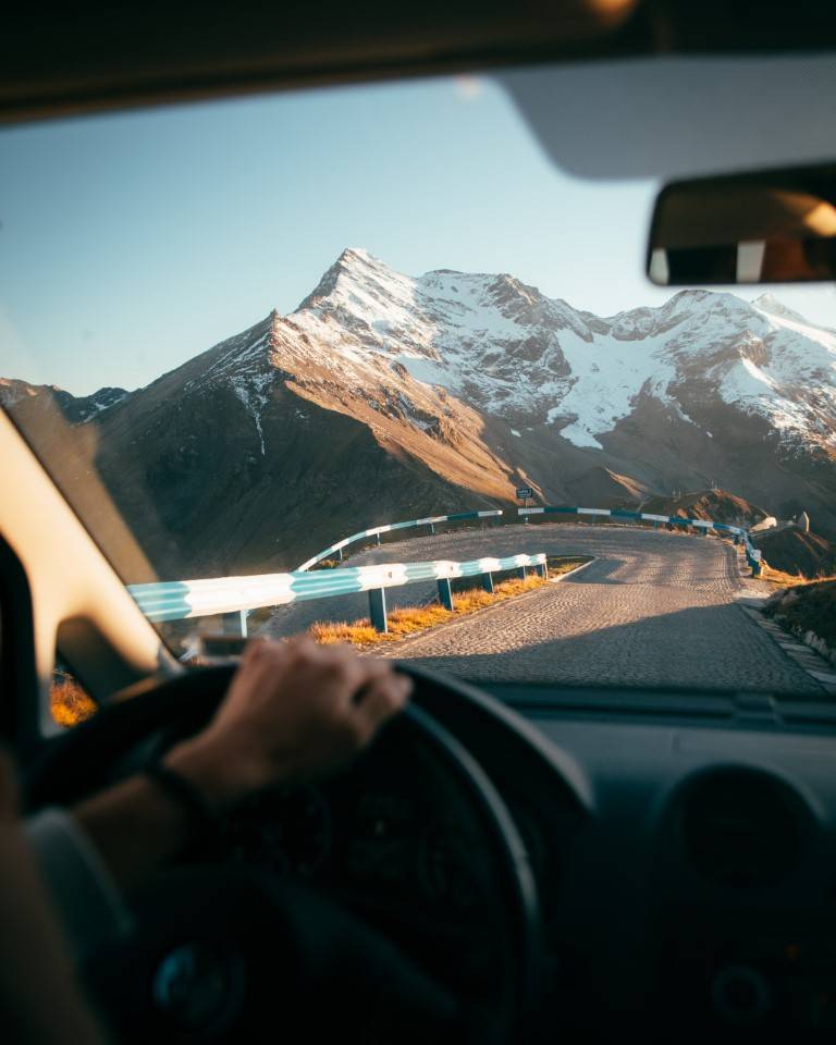 View from the driver's seat past the steering wheel to a mountain road and a snow-covered peak