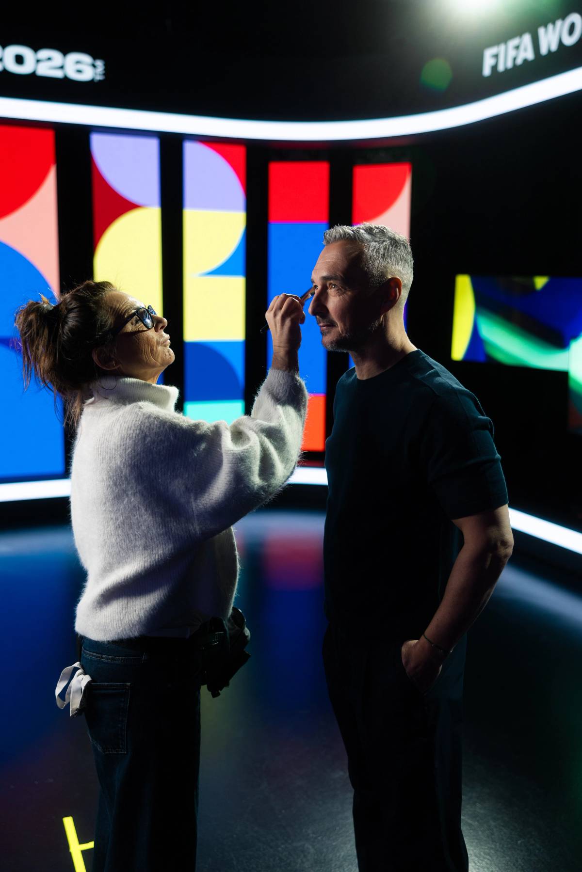 A makeup artist applies makeup to a man on a studio set with colorful illuminated panels.